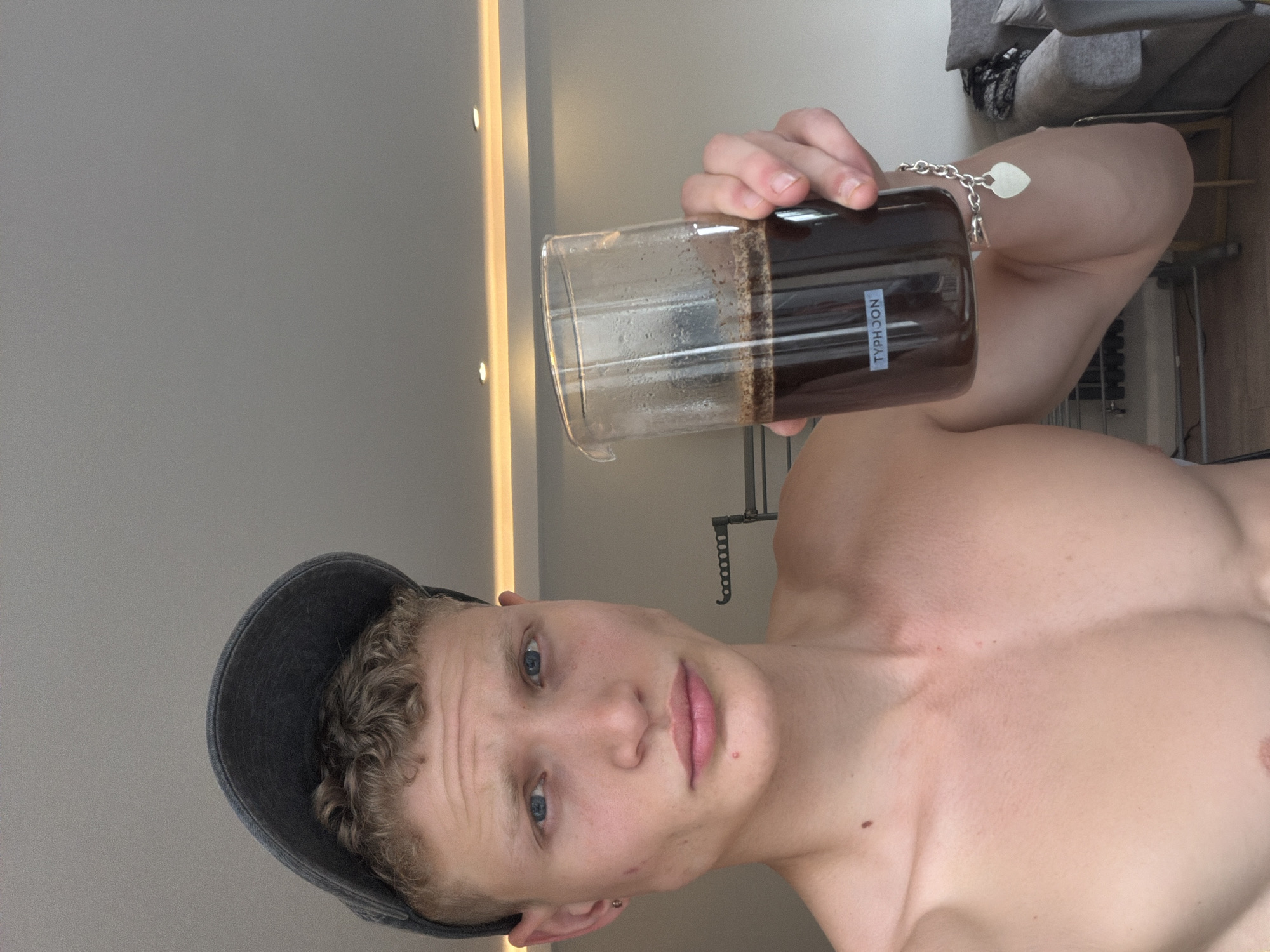 A young, fair-skinned male with blue eyes and light brown curly hair is pictured from the chest up, wearing a dark baseball cap and holding a glass of dark liquid, possibly coffee, in his right hand. He is shirtless and appears to be indoors, with a ceiling with recessed lighting visible in the background. A faint reflection suggests a window or light source to the right. The scene is a domestic indoor setting, consistent with an apartment or house. No specific landmarks or indicators of a London location are present.