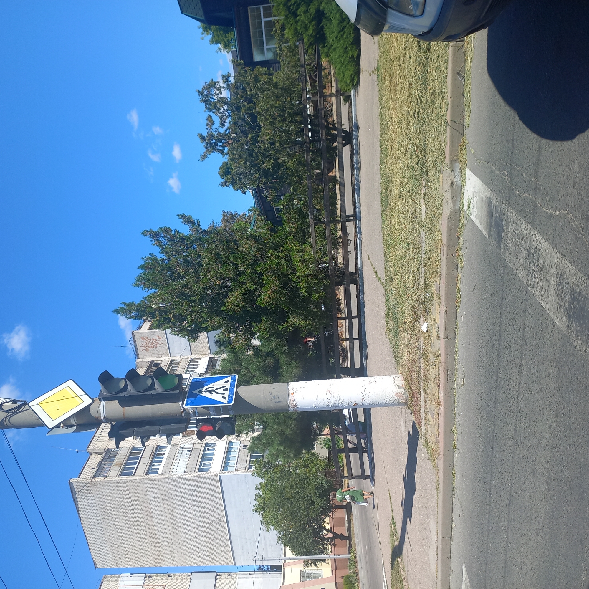 The image depicts a street scene in Kropyvnytskyi, Ukraine, under a clear blue sky. In the foreground, a pedestrian crossing is visible on the asphalt road. To the right, the front of a light-colored vehicle is partially in view, with its headlight and bumper visible. A dark shadow of the vehicle is cast on the road.
A white concrete pole with peeling paint stands prominently on the right side of the image, serving as a support for traffic signals and signs. A pedestrian crossing sign, a blue square with a white silhouette of a person walking, is attached to the pole. Above it, a traffic light shows a red signal on the top and a green signal on the bottom. A yellow diamond-shaped sign with a black border is mounted above the traffic lights.
Further back, a woman in a green dress with a white bag slung over her shoulder is walking away from the camera, crossing the street. Behind her, a row of trees forms a green barrier, with a wooden fence running horizontally behind the trees. In the background, multi-story apartment buildings with light-colored brick facades are visible, one of which has a patterned decorative element on its upper section. The environment suggests an urban setting with residential buildings, a road, and green spaces.