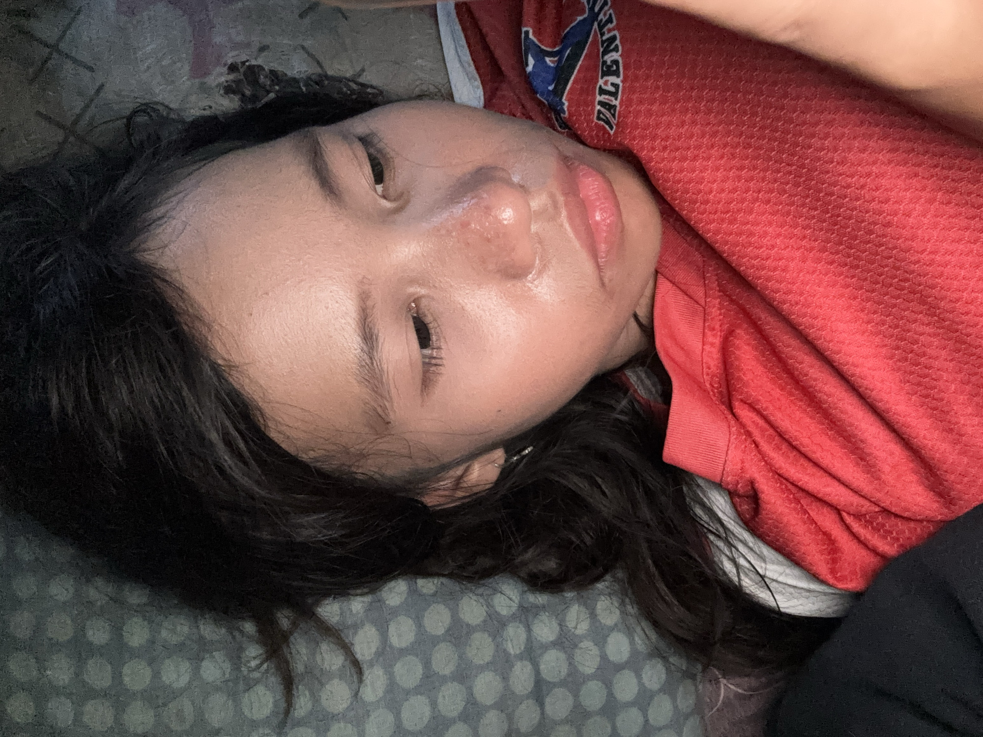 The media file is a close-up, horizontally oriented photograph of a young person lying down, with their head tilted back and their eyes partially open. They have dark, long hair that frames their face. The person is wearing a red shirt with white and blue lettering visible on the collar area, which appears to be a sports jersey. The background consists of a gray and white polka-dot patterned fabric, possibly a blanket or bedsheet, and a darker, textured surface.
The lighting in the image suggests it was taken indoors, potentially in the evening or with artificial lighting, as there are strong highlights on the person's skin, indicating a glossy or slightly oily complexion. The person's expression is neutral, perhaps a bit tired or relaxed. There are no obvious clues about the weather. The location context indicates East Jakarta, Indonesia, but there are no specific visual cues within the image to confirm this. The overall scene is intimate and personal, focusing on the subject's face and upper body.