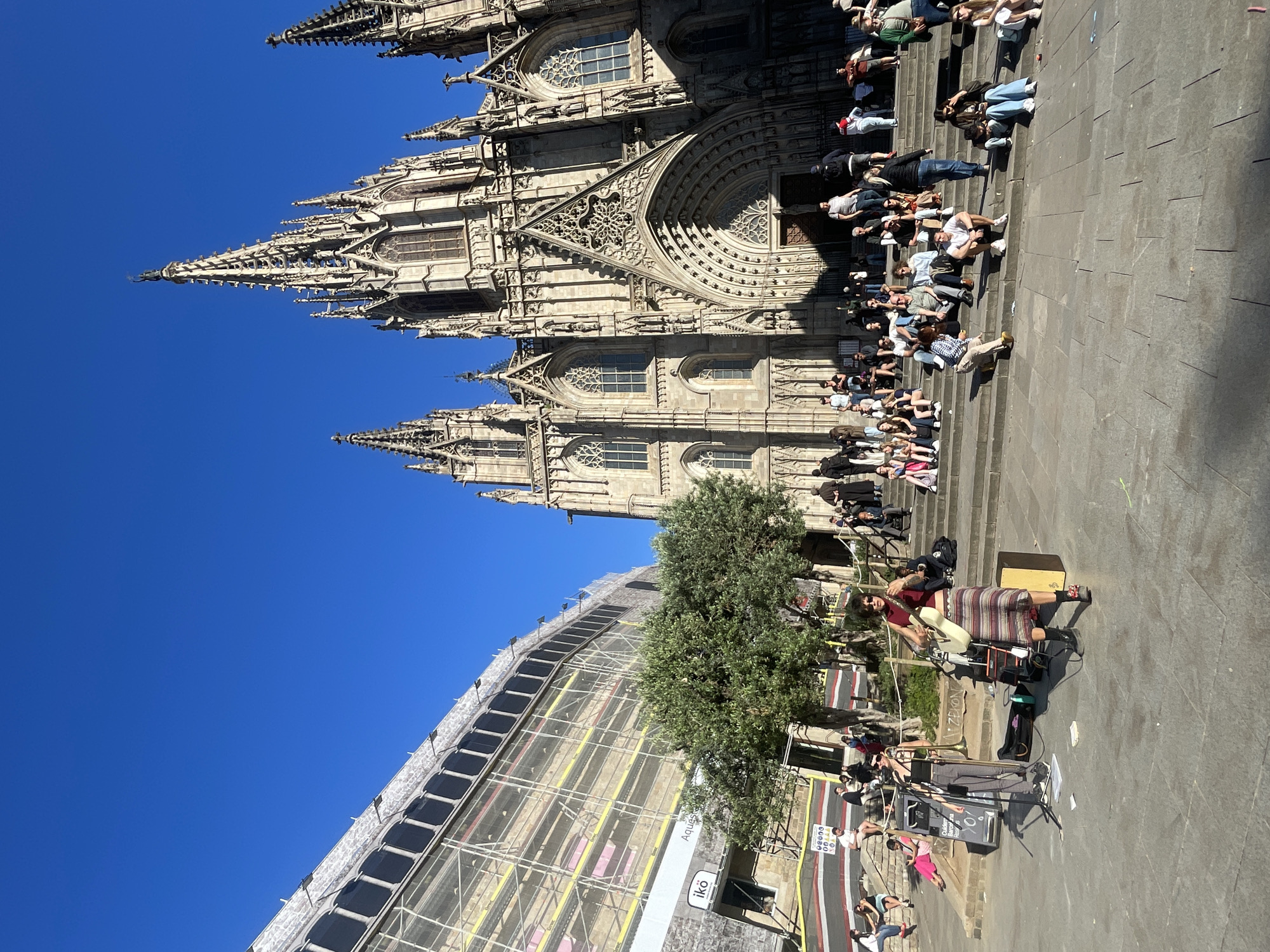 The image shows a scene in Barcelona, Spain, featuring the Barcelona Cathedral. The cathedral, a large Gothic structure, dominates the background. Many people are sitting on the steps leading up to the cathedral. In the foreground, a musician plays the saxophone while seated on a wooden stool, with sound equipment nearby. A tree partially obscures the view of the building to the right of the cathedral, which is under construction and covered in scaffolding. The sky is clear and blue.