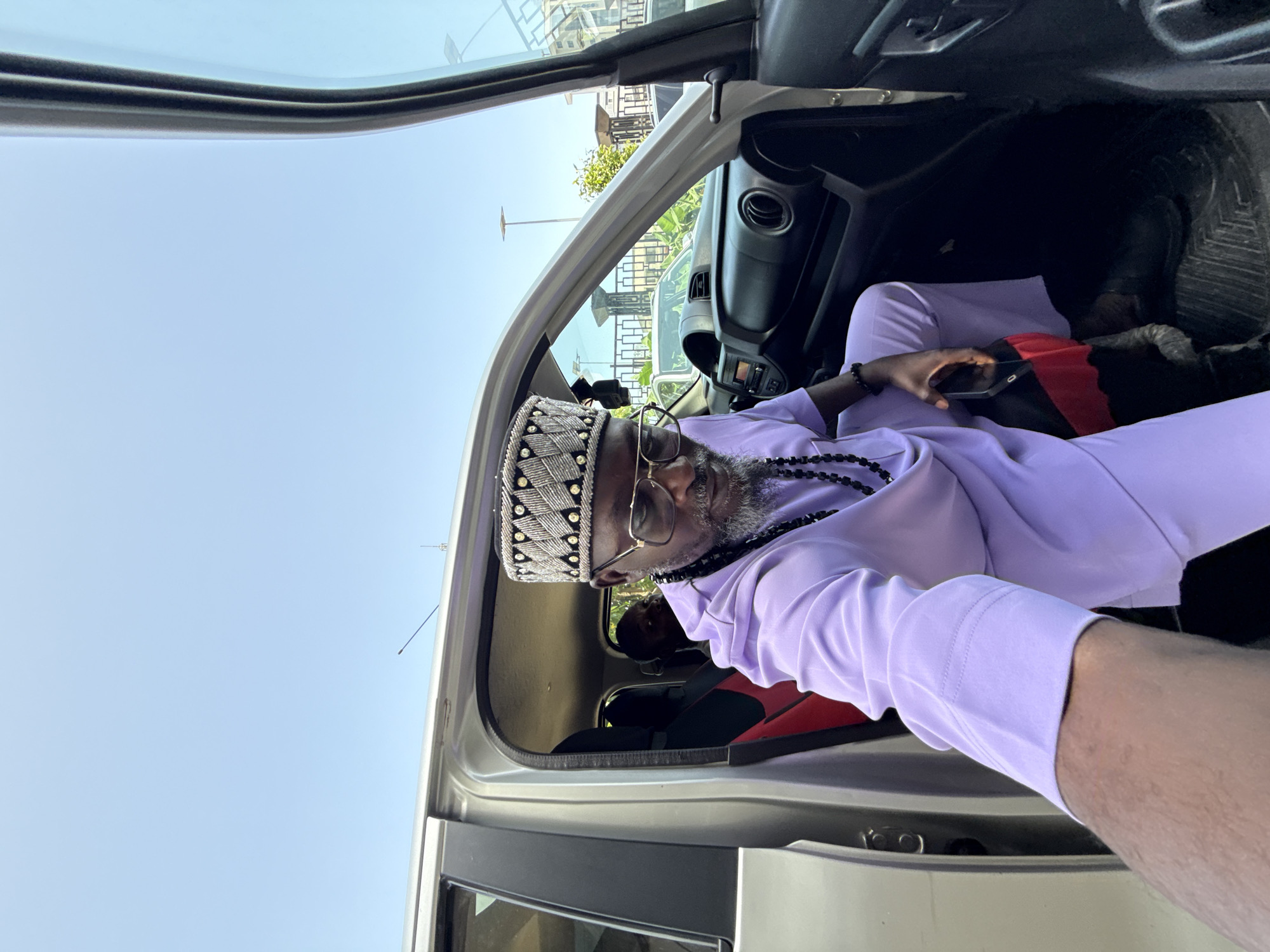 An image of a man with grey beard sitting inside a vehicle, possibly a car, with the door open. He is wearing glasses, a beaded cap, a light purple long-sleeved shirt, and a beaded necklace. He is holding a phone in his hand. The environment shows a partly sunny outdoor scene that suggests the location is outside a building, possibly in a parking lot. The specific location within Itirin, Nigeria, cannot be determined.