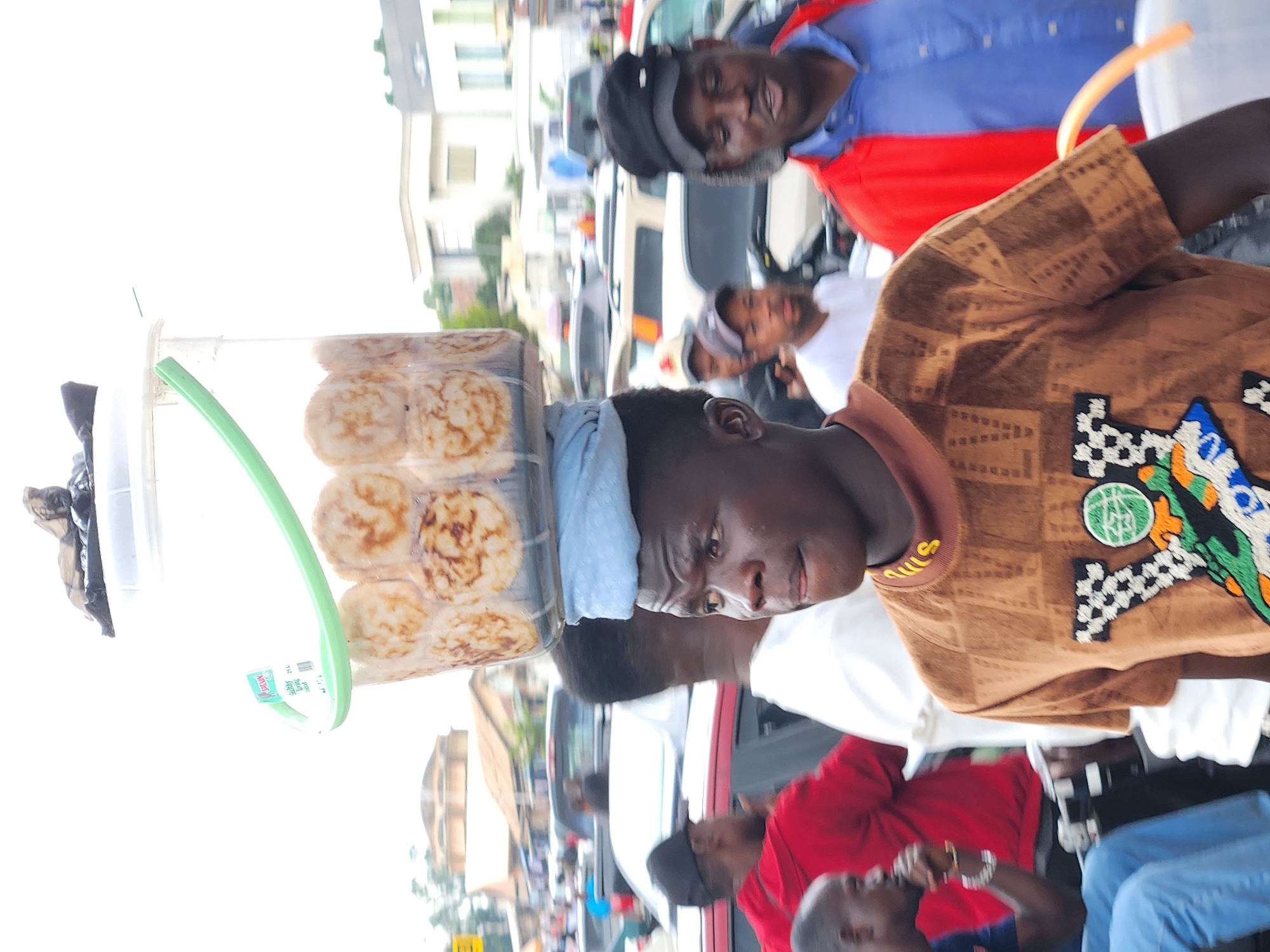 The image shows a street scene with several people. In the foreground, a young man wearing a light blue headwrap and a brown patterned shirt is looking to the right. He is holding a clear plastic container filled with round, flat food items that appear to be pancakes or fritters. A green plastic tube extends from the container. Behind him, other individuals are visible, including a person in a red shirt and a person in a blue and red jacket. Some of the people are wearing hats. The background consists of buildings and vehicles, suggesting an outdoor urban or market setting. The lighting is bright, indicating it might be daytime. There is some text visible on a sticker on the plastic container, which appears to read "Sammy" and "21L". The overall atmosphere seems bustling, possibly a market or street food vendor scene. The orientation of the image is sideways.