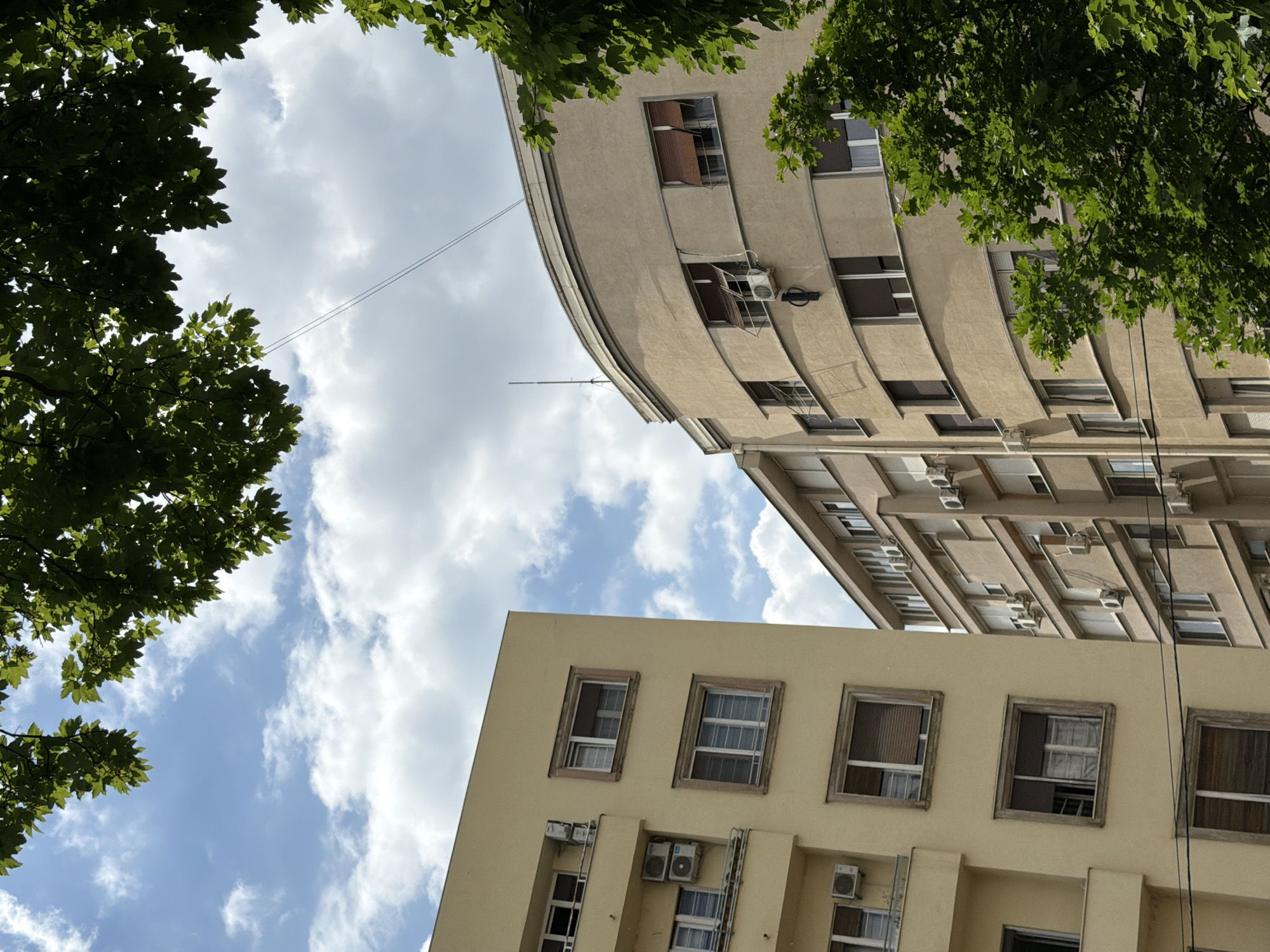 The image depicts an upward view of two buildings in Belgrade, Serbia, framed by tree foliage against a cloudy sky. The building on the right is a tall, curved structure with multiple floors and numerous windows, many equipped with air conditioning units. The building on the left is a more rectangular structure, also with multiple floors, featuring windows and several air conditioning units attached to the exterior. An electrical wire is visible stretching across the sky between the trees and the curved building.