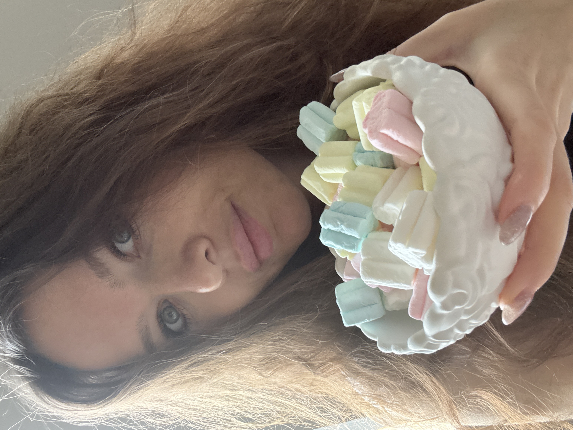 A woman with long brown hair is holding a white ceramic container filled with pastel-colored marshmallows. Her face is visible, with a slight smile and light-colored eyes. The background is a blurred white. The location is not discernible from the image, so I cannot provide information about Khimki, Russia.