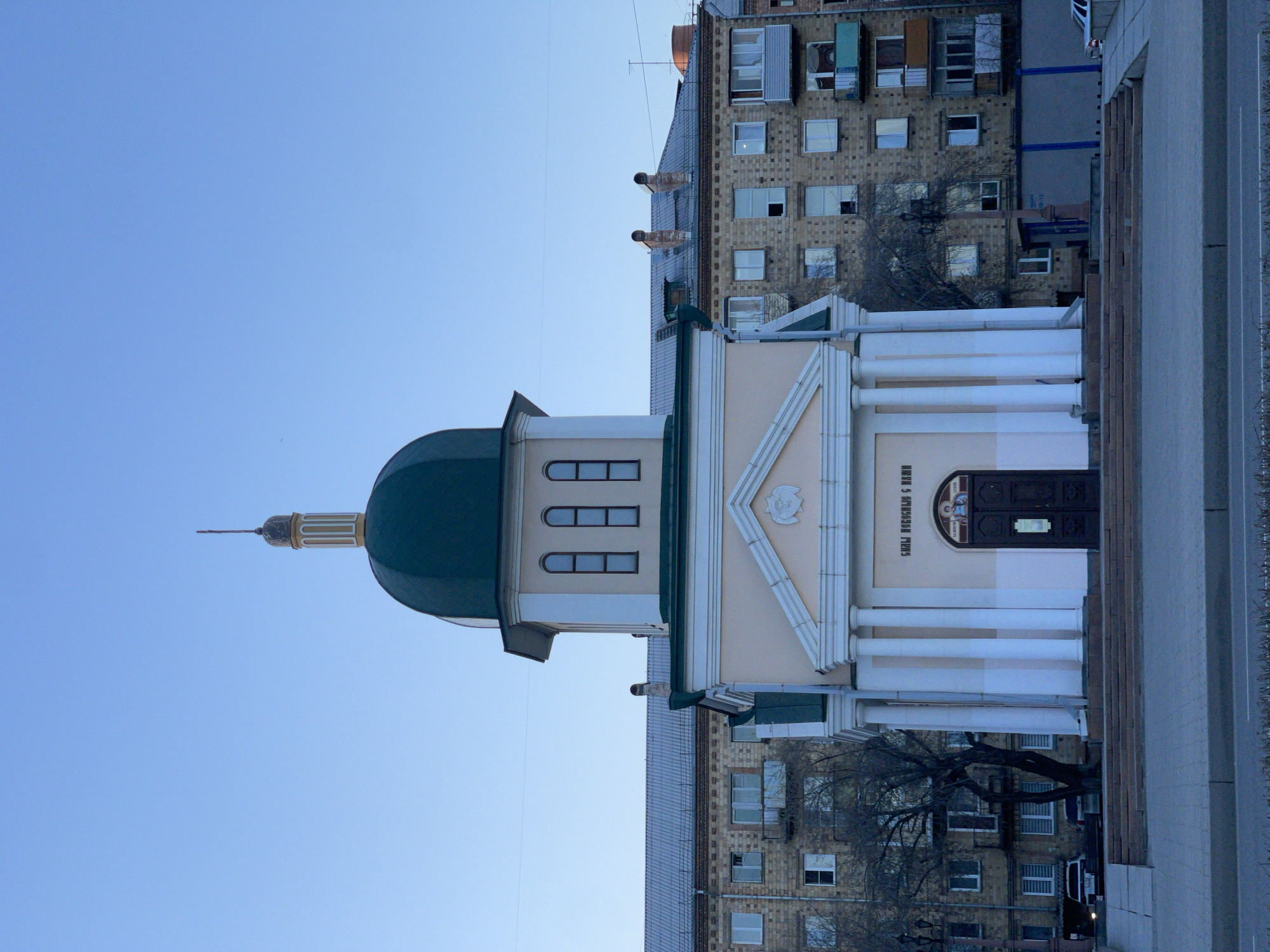 An urban landscape in Krasnoyarsk, Russia, is captured under a clear, bright blue sky. The central focus is a light-colored religious building, likely a chapel or small church, distinguished by its dark green dome and a slender, ornate steeple culminating in a spire. This structure features a classical-style facade with a prominent portico, comprised of white columns supporting a triangular pediment. The pediment is adorned with a decorative, carved relief.
Above the dark, arched entrance door of the religious building, Cyrillic text reads "СВЯЩ. ИОСИФА ОБРУЧНИКА," which translates to "Holy Joseph the Betrothed." An icon is also visible above the doorway. To the right and behind this building stands a multi-story apartment block constructed of light-colored brick, characterized by numerous windows of varying sizes, some with small balconies or ventilation units. A bare deciduous tree is positioned between the two buildings, its branches reaching towards the apartment block. In the immediate foreground, a paved area, possibly a sidewalk or plaza, runs parallel to the religious building.
The scene appears to be set during daytime, with soft, even lighting suggesting either morning or late afternoon. The sky is completely cloudless, indicating fair weather. No people are visible, and no specific activities are taking place, contributing to a sense of stillness in the urban environment.