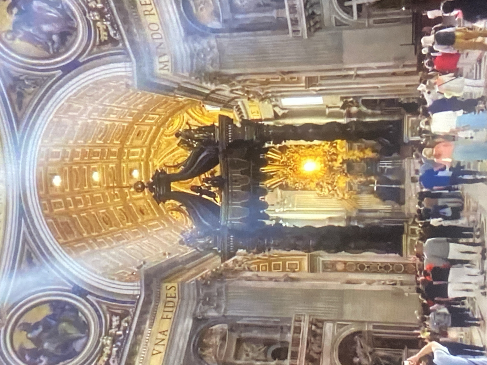 The image depicts the interior of a grand basilica, likely St. Peter's Basilica in Vatican City, not Tinaquillo, Venezuela. The view is angled upward towards the ornately decorated dome and ceiling. The Baldachin, a large bronze canopy, is a central feature, with its dark, twisted columns and golden accents. In the background, there's a glimpse of the altar area with a radiant light source. Rows of people, presumably tourists, line the edges of the scene, observing the basilica's architecture. The image captures the scale and artistry of the religious structure.