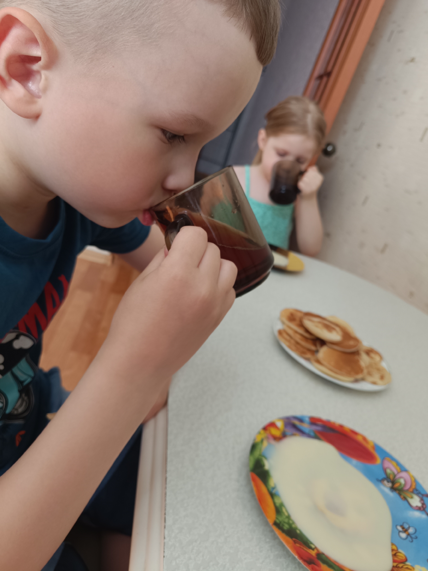 Two children are at a table with food and drinks. A boy in a blue shirt is drinking from a glass, while a girl in a teal tank top is drinking from a glass in the background. There is a plate of pancakes and a plate with white substance (likely cream or sour cream) on the table. The setting appears to be a domestic kitchen or dining area. It is not possible to determine whether this is in Tobolsk, Russia, or any other specific city without additional information.