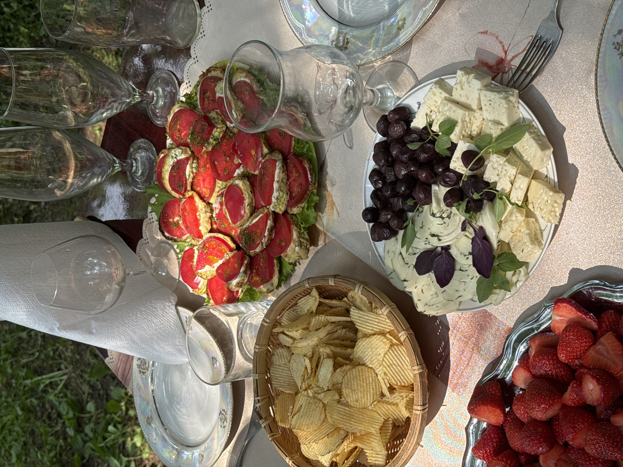 This image depicts an outdoor picnic table set with various food items and glassware. The setting appears to be in a garden or park area, with greenery visible in the background.
On the table, there is a platter of what appears to be appetizers made with sliced tomatoes and a creamy filling, garnished with herbs. Adjacent to this, a woven basket is filled with ridged potato chips. A separate plate displays a selection of cheeses cut into cubes and slices, accompanied by a pile of dark olives and fresh basil leaves. A silver platter on the left is filled with halved strawberries.
Several empty wine glasses and a clear glass tumbler are positioned around the table. A white, textured napkin is also visible. The table is covered with a patterned tablecloth. The lighting suggests it is daytime, with sunlight casting shadows. There are no people visible in the image. The picnic appears to be set up for a gathering, but no individuals are present in the frame. The location is specified as "Козино, Russia" in the prompt, implying this is a picnic in that geographic area.