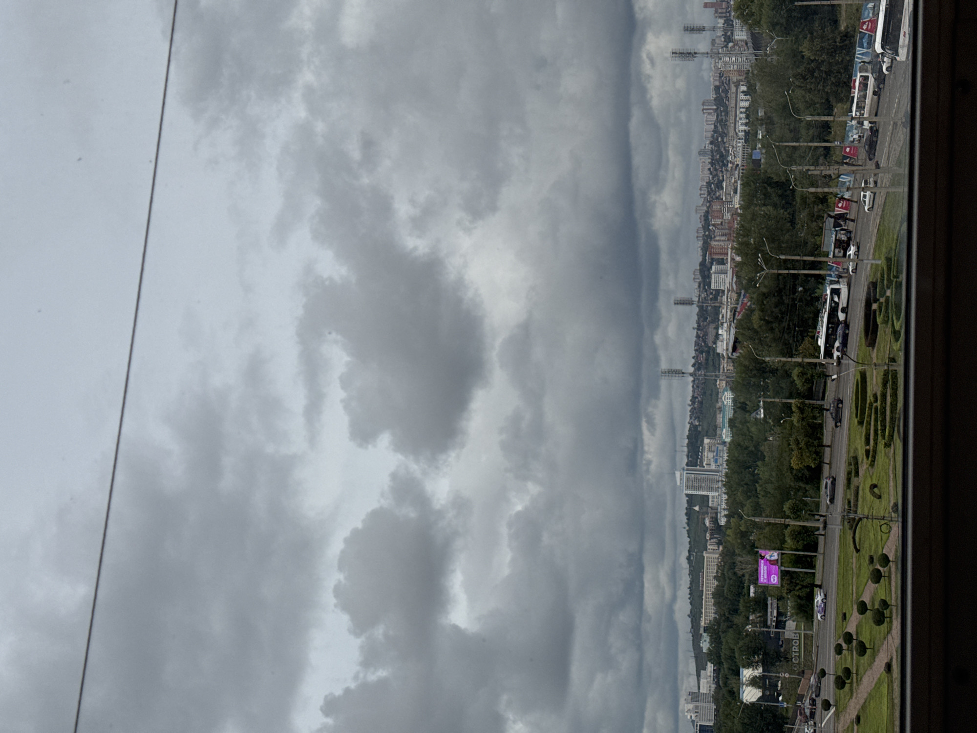The image shows a cloudy, overcast sky over a cityscape. In the foreground, a manicured park with topiaries and hedges is visible. Beyond the park, a multi-lane road is lined with trees and streetlights. Several vehicles, including buses and cars, are traveling on the road. In the background, a dense urban area with various buildings, including skyscrapers and residential structures, stretches across the horizon. Several tall radio towers are also present. The scene appears to be taken from an elevated vantage point, possibly a building overlooking the city. A thin wire is visible across the top of the image. The overall atmosphere is somber due to the cloudy sky. The sign that says "ОСТРОВ" (Ostrov, meaning "Island" in Russian) on the left side of the road indicates the location is Ostrovsky Island in Krasnoyarsk, Russia.
