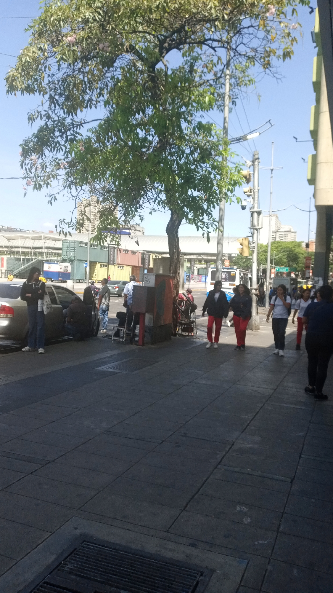 The image depicts a city street scene. Several people are walking on the sidewalk, and a group is gathered around a parked car on the left side of the frame. There is a tree providing shade, and a traffic light post is visible. In the background, there are cargo containers and a building, suggesting an urban or commercial area. The sky is clear.