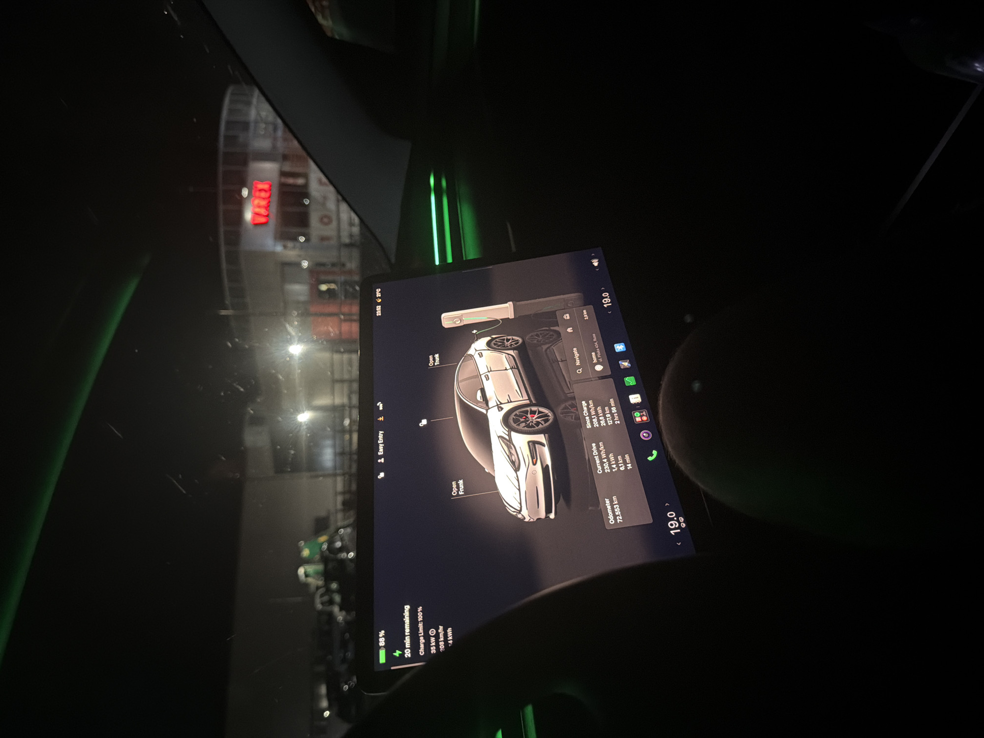 The image shows the interior of a Tesla electric vehicle at night, likely in Ruse, Bulgaria. The car's large touchscreen display is prominent, showing a charging status with 88% battery remaining and an estimated 20 minutes until fully charged. The display also features a 3D model of the car, indicating the open trunk and a charging station. Vehicle statistics such as odometer reading (72,553 km), current drive efficiency, and since charge statistics are visible. Navigation information suggests a destination named "home" at "ul. Plovdiv 434, Ruse".
Outside the vehicle, through the windshield, a building with a lit sign that reads "VIREX" is visible. The surrounding area appears to be an outdoor parking lot or charging station, with other vehicles and infrastructure dimly illuminated in the darkness. Green ambient lighting is present inside the car's cabin, reflecting on the interior surfaces. The time is displayed as 23:52, with an outside temperature of 21°C.