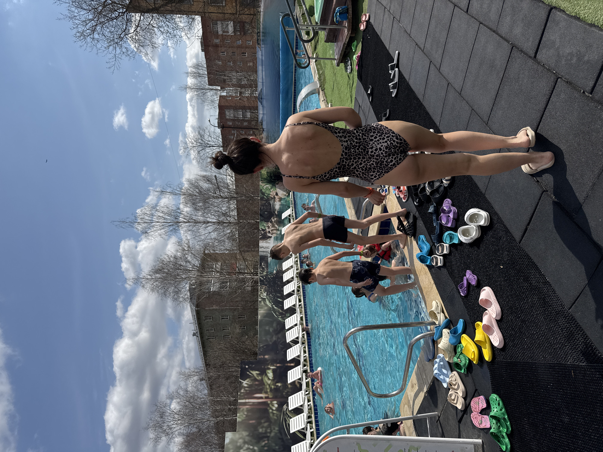 The image shows an outdoor swimming pool in Votkinsk, Russia. A woman in a leopard-print swimsuit stands at the edge of the pool. Two boys are about to enter the pool. People swim in the pool. Many pairs of sandals and flip-flops lie on the black rubber pavement near the pool's edge. A building is visible in the background. The sky is blue with clouds, and there are trees without leaves.
