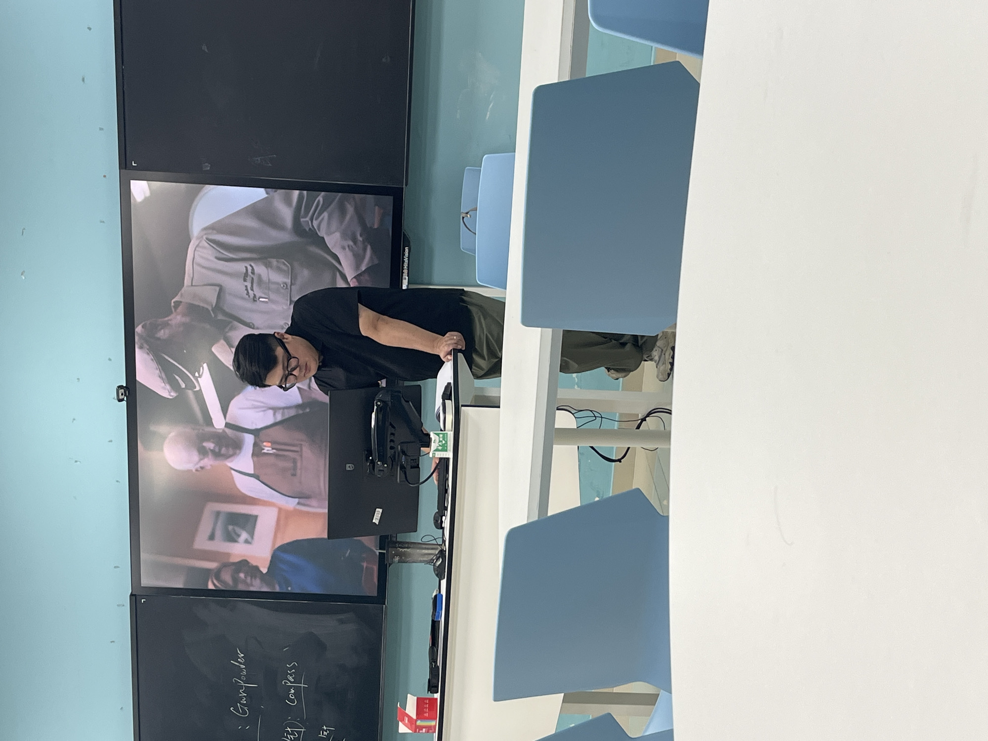In a classroom setting, likely at a school in Jiangjin, China, a young Asian man wearing glasses and a black t-shirt stands behind a white table, looking down at a stack of papers. A large monitor displays a paused video of three people, two men and one woman, seemingly engaged in a demonstration or discussion. One of the men on the screen wears an apron. The room features light blue walls, a black whiteboard with some handwritten notes, and several light blue chairs arranged around white tables. The overall impression is that of an educational environment where a presentation or lecture is taking place.