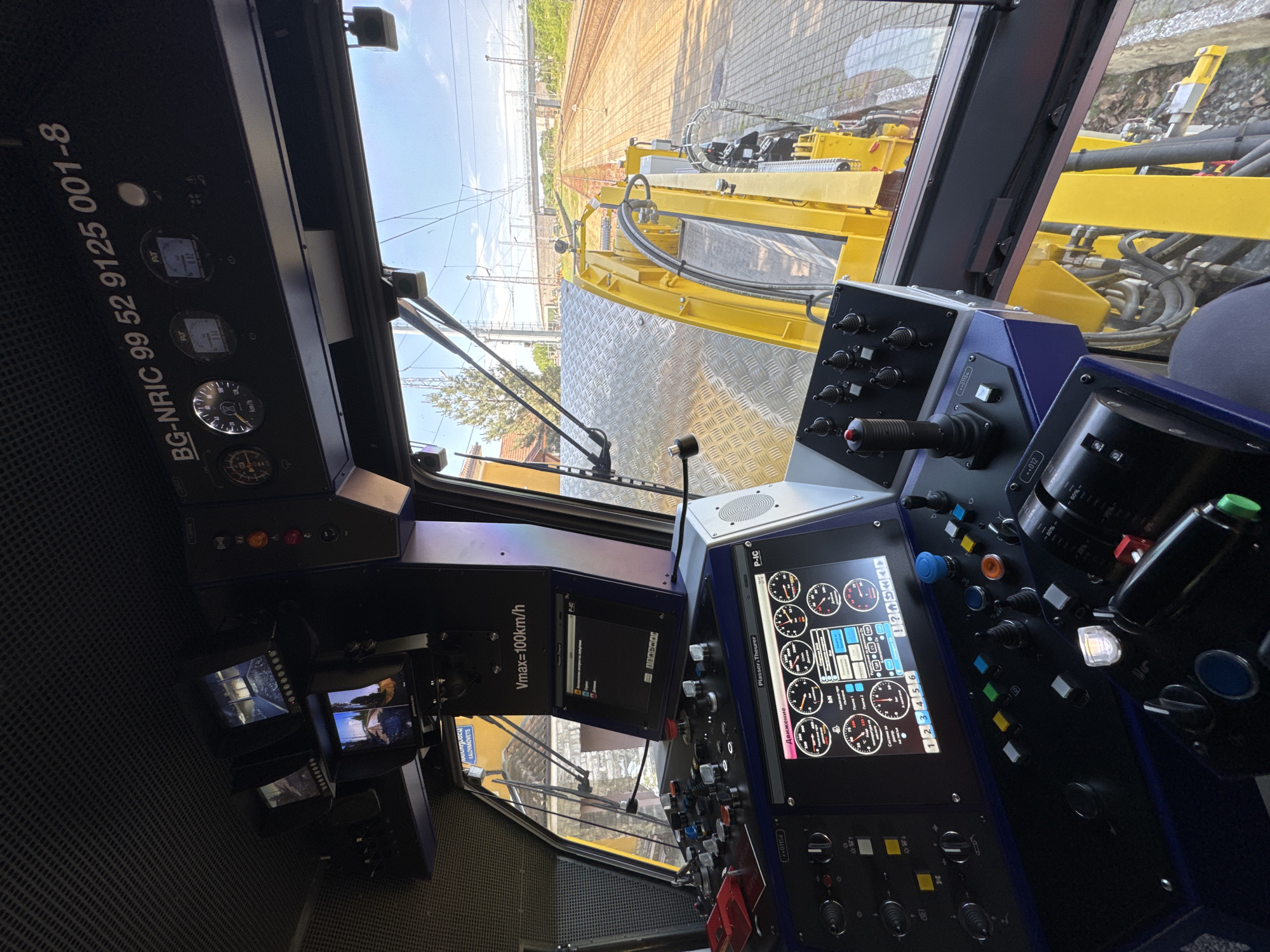 This is a view from inside the cabin of a specialized railway maintenance vehicle or machine. The perspective is looking forward through the windshield, showing the track ahead and some of the machinery attached to the vehicle, specifically a yellow, track-related apparatus.
Inside the cabin, there are several control panels with various buttons, switches, gauges, and displays. One display shows gauges and indicators labelled "Движение" (Movement). Another screen indicates "Vmax=100km/h". Text labels on the panels are in Bulgarian and English. On the left panel is "BG-NRIC 99 52 9125 001-8". Also, the brand name "Plasser & Theurer" is shown on the screen. Other screens show video feeds, presumably from cameras mounted on the vehicle. The city of Kaloyanovets, Bulgaria is mentioned on one of the video screens.
