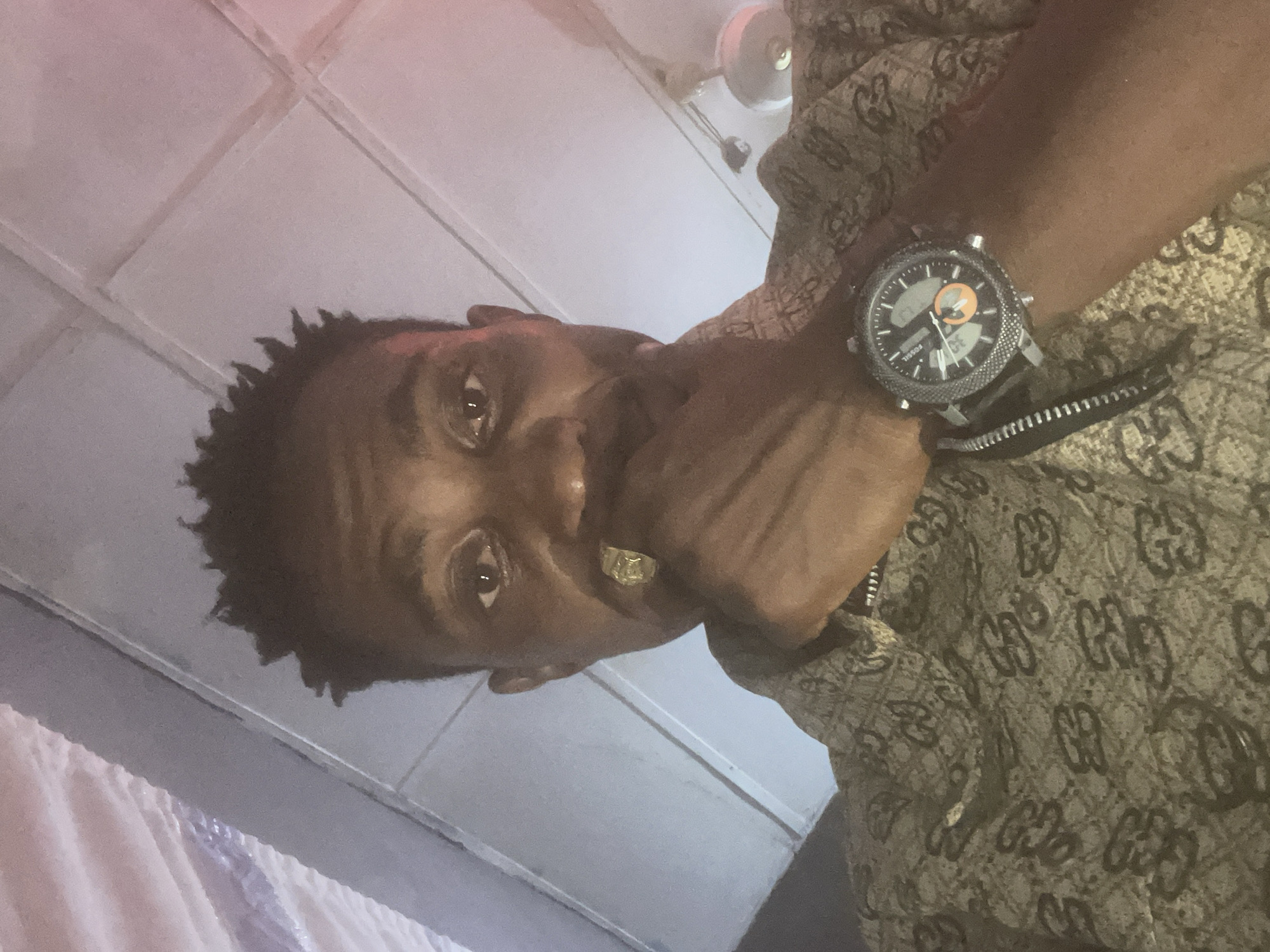 In Adankolo, Nigeria, a man with dark skin and short, spiky hair takes a low-angle selfie. He is wearing a light brown shirt with a repeating dark brown pattern. He is holding his right hand to his mouth, and a gold ring is visible on his right ring finger. On his left wrist, he wears a dark-colored wristwatch with a circular face, displaying an analog and digital time. The background appears to be an interior with a drop ceiling, and a light fixture is visible in the upper right corner.