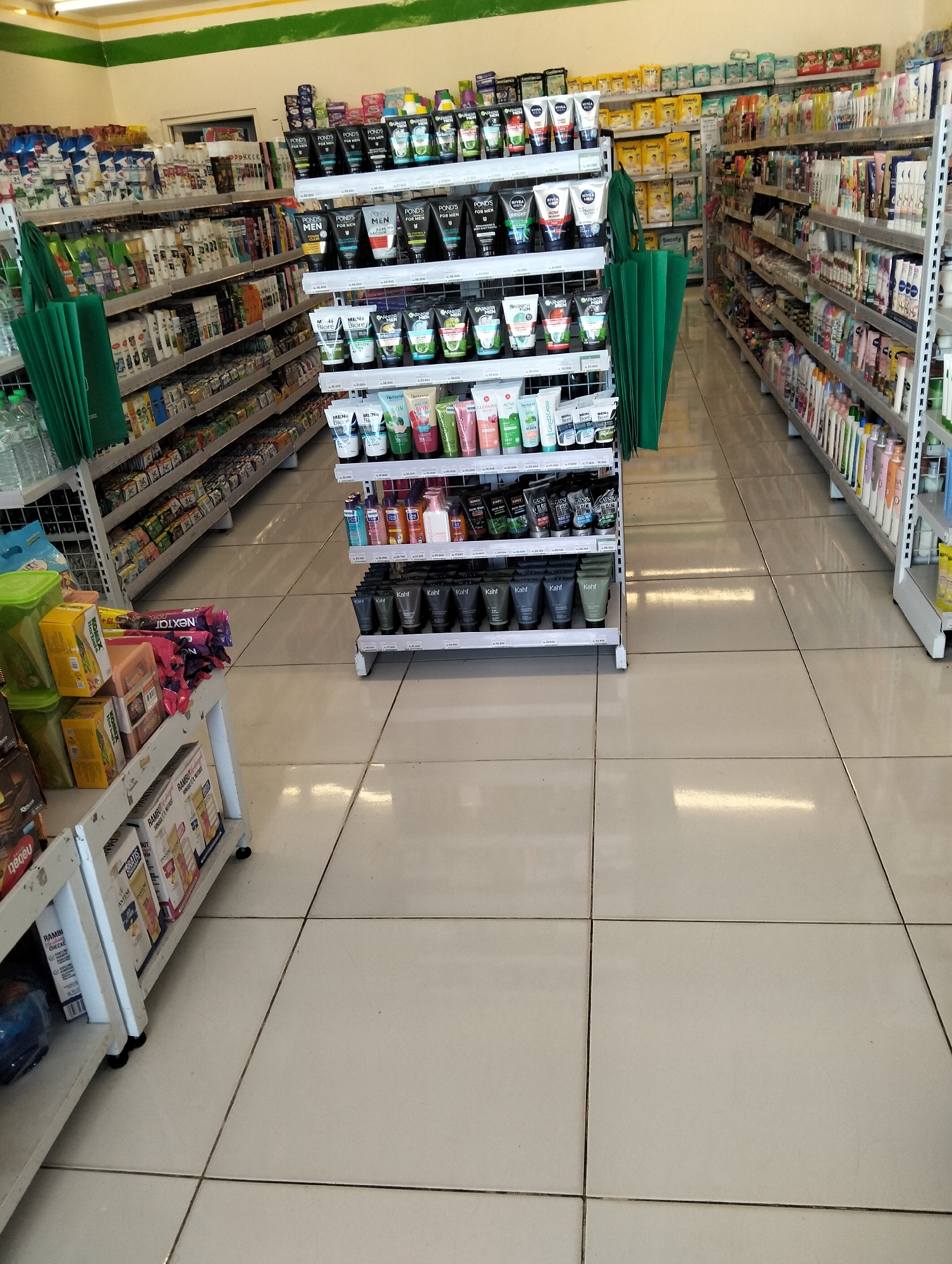 The image shows the interior of a small convenience store in Muncar, Indonesia. The store is filled with shelves stocked with various consumer goods, including personal care products, food items, and drinks. The flooring is tiled, and fluorescent lighting illuminates the scene. In the foreground, there are several shelves displaying personal care products like lotions and shampoos. The shelves extend to the background, with more products lining the walls. The overall impression is of a well-organized retail space typical of a convenience store.