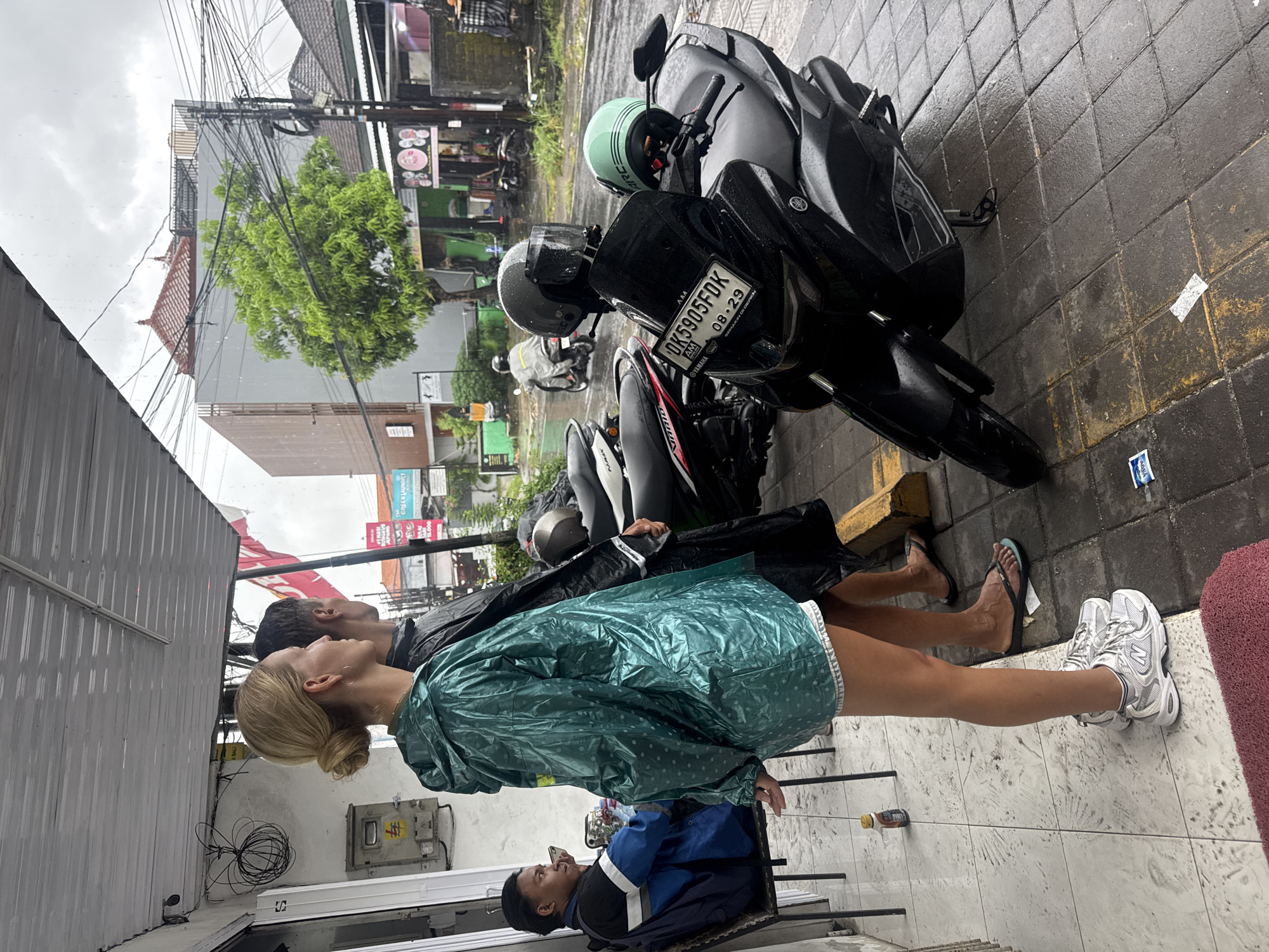 The image captures an urban street scene in Kuta Selatan, Indonesia, on an overcast and rainy day. The ground, including the sidewalk and road, appears wet.
In the foreground, two individuals, a man and a woman, stand side-by-side, facing away from the camera and looking upwards or straight ahead. The woman, with blonde hair tied back, wears a teal-colored, polka-dotted raincoat over light-colored shorts and white sneakers. The man, with dark hair, wears a dark jacket. Further back and to the right, another man is seated in a chair, wearing a blue and white uniform jacket.
Several motorbikes are parked along the wet sidewalk. One prominent black motorbike in the middle ground has a visible license plate that reads "DK 5905FDK" and "08-29", with "YAMAHA" and "SAMANJU" text below. Two helmets, one light green and one black, rest on the motorbikes.
The background features various buildings with typical Indonesian architecture, including reddish-brown tiled roofs. Numerous electrical wires crisscross overhead. In the distance, a commercial sign for "GREEN LAUNDRY" is visible, with Indonesian words like "MURAH" (cheap) and "TERMURAH" (cheapest), along with prices "Rp 15.000" and "Rp 10.000". A small blue and white packet lies on the wet tiled ground near the motorbikes. The overall impression is a bustling street during a downpour or immediately after.