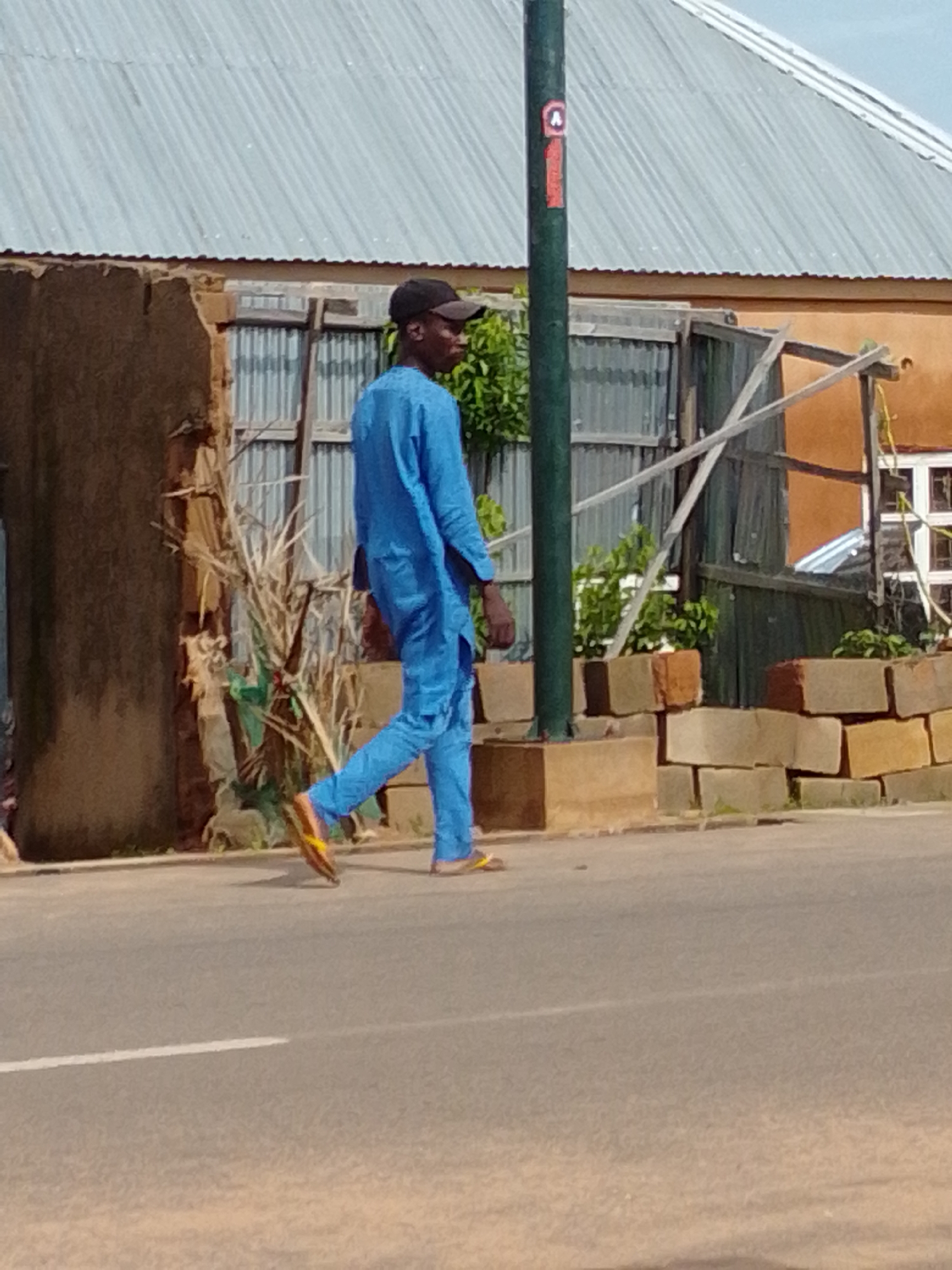 In Gombe, Nigeria, a man walks on a paved road. He is wearing a blue kaftan and matching pants, along with yellow and brown flip-flops. A black baseball cap covers his head. The man is positioned to the right of a dark green, cylindrical pole which has a red and white sticker on it. Behind the man and the pole is a building with a corrugated metal roof. The building has a light brown facade and a partially constructed wall made of concrete blocks. The background includes vegetation and a wooden structure. The road has a white line painted down its center.