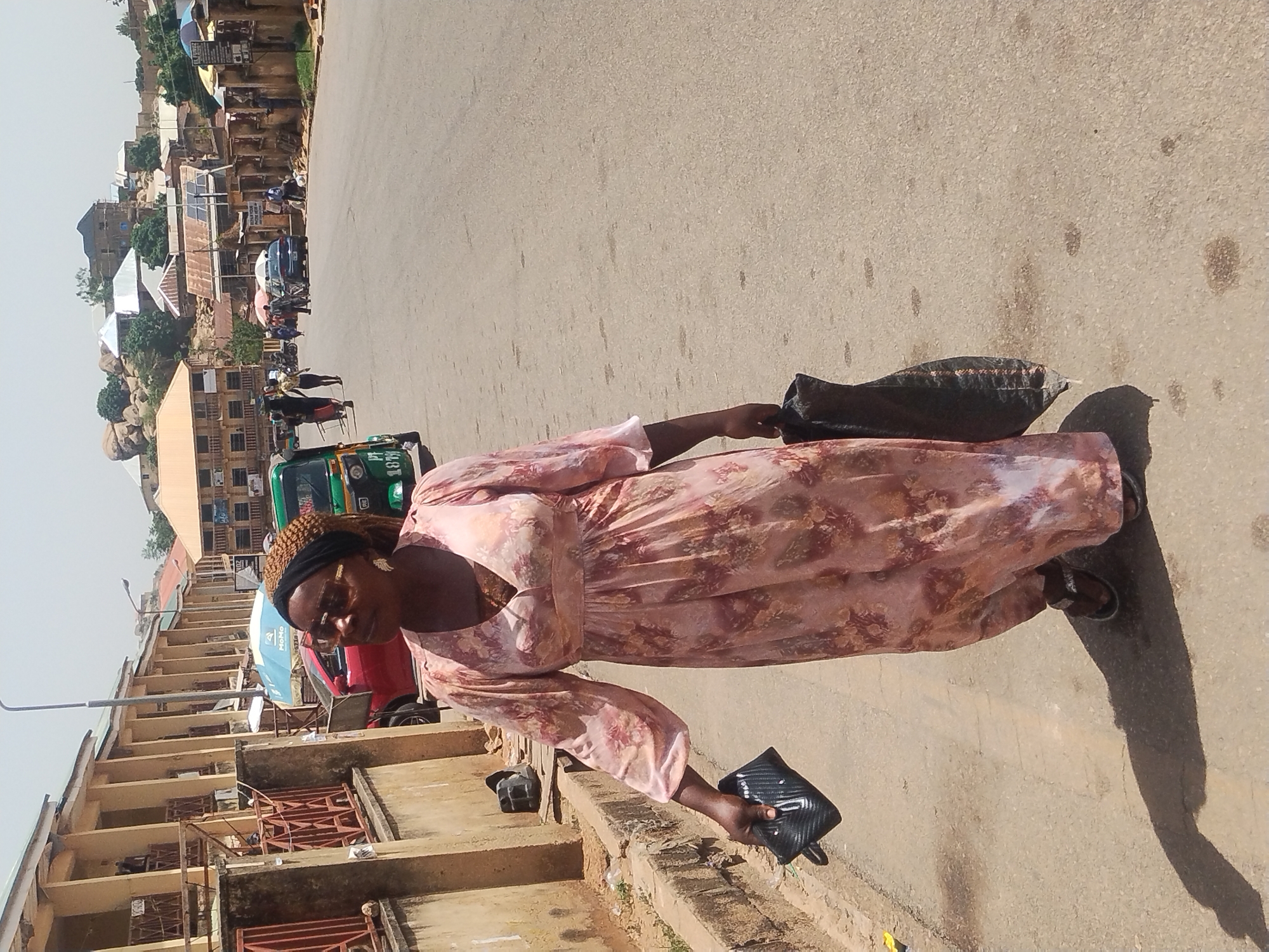 An eye-level outdoor shot depicts a woman walking on a paved street in Jos, Nigeria. She is wearing a long, floral-patterned dress and sandals, holding a black clutch in one hand and a black tote bag in the other. Her head is adorned with a black headband and eyeglasses. The street exhibits light stains and imperfections on the asphalt surface. Behind her is a row of multi-story buildings featuring orange brick facades, concrete balconies, and iron railings. In the background, there's a mix of vehicles, including a green tricycle. Higher in the background, a series of residential buildings and businesses are situated on an elevated plane with visible rocks and trees. The lighting suggests a bright, sunny day, casting shadows of the woman and objects in the scene.