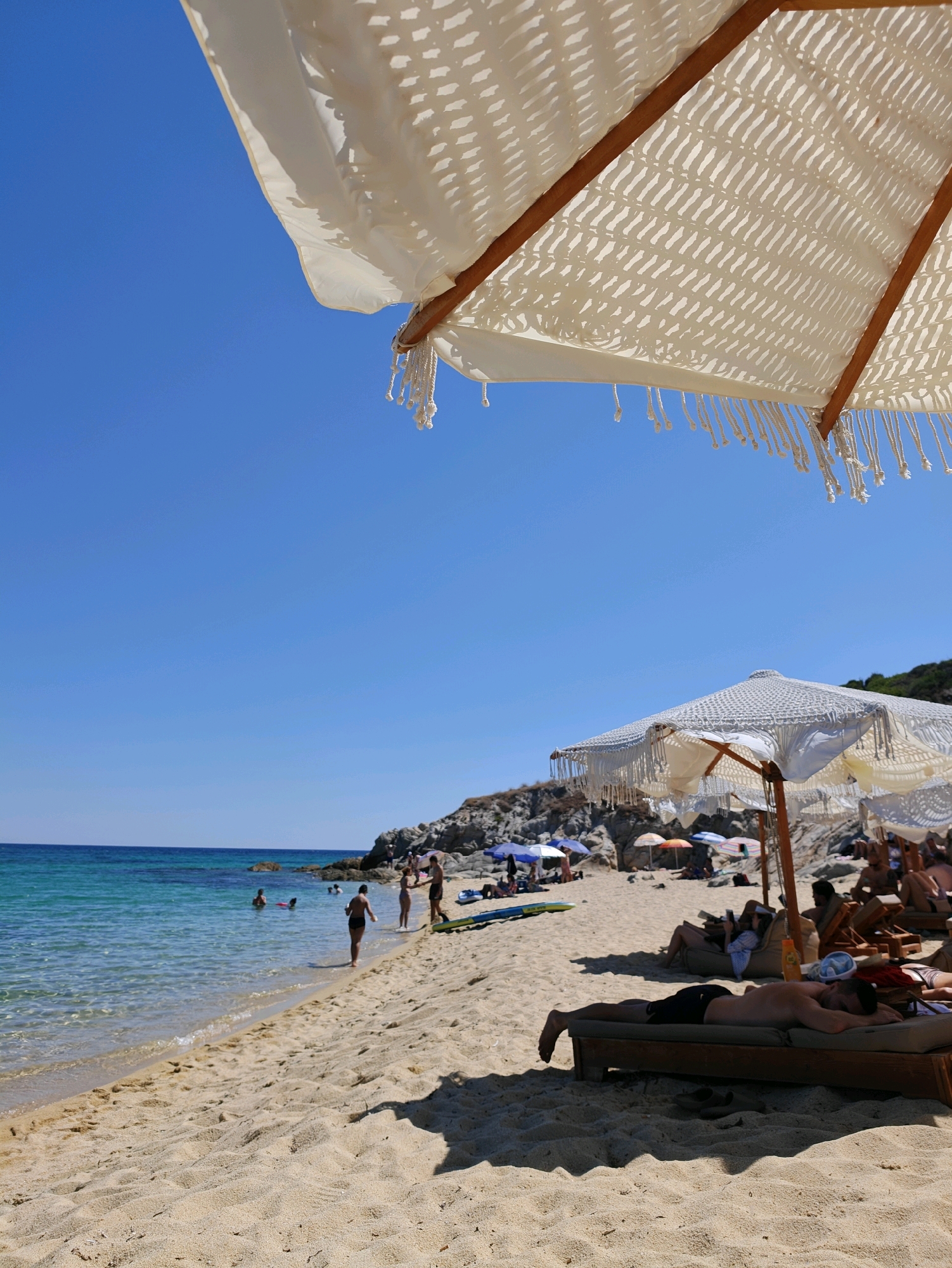 In the Municipal Unit of Toroni, Greece, a sandy beach features a person lying face down on a lounge chair under a large, white, macrame-style umbrella with tasseled edges. The umbrella is in the foreground, angled towards the sky, casting a shadow on the sand. Beyond the lounge chair, other beach umbrellas and loungers are visible, some occupied by people. Several individuals are seen walking along the shoreline, wading in the clear, turquoise water, or swimming. The sea extends to the horizon under a bright blue sky. Rocky formations with some vegetation are present along the coastline. The sand is textured with small ripples and shadows.