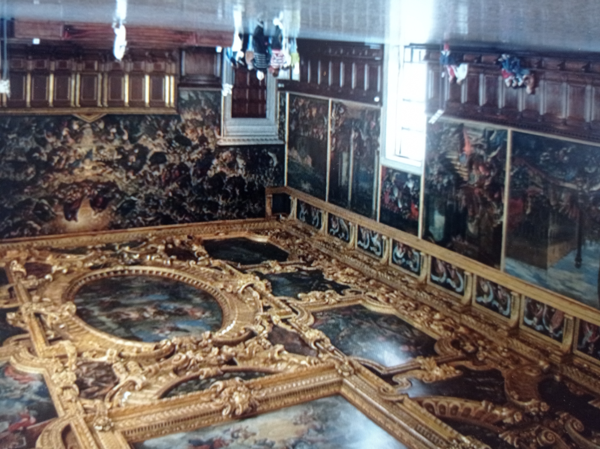 This image depicts the interior of a grand hall, likely a palace or museum, with ornate gold-leaf detailing and large-scale paintings adorning the walls and ceiling. The paintings on the ceiling feature mythological or religious scenes with multiple figures, rendered in rich colors. The walls are lined with a series of framed paintings, some depicting architectural scenes and others with numerous figures engaged in various activities. Below the paintings, dark wooden paneling runs along the lower section of the walls, with benches situated in front of it.
Several individuals are visible in the hall. In the foreground, on the left, a group of people are seated on a long wooden bench. Further into the hall, a group of people are standing and walking. One person in a white garment is walking towards the right side of the frame, near a set of tiered seating. The floor is reflective and appears to be made of polished stone or marble.
The lighting in the hall is a mix of ambient light, likely from windows or overhead fixtures, and possibly some directed lighting highlighting the artworks. The overall impression is one of opulence and historical significance. The image does not contain any discernible elements that would indicate it is located in the city of Mukachevo, Ukraine.