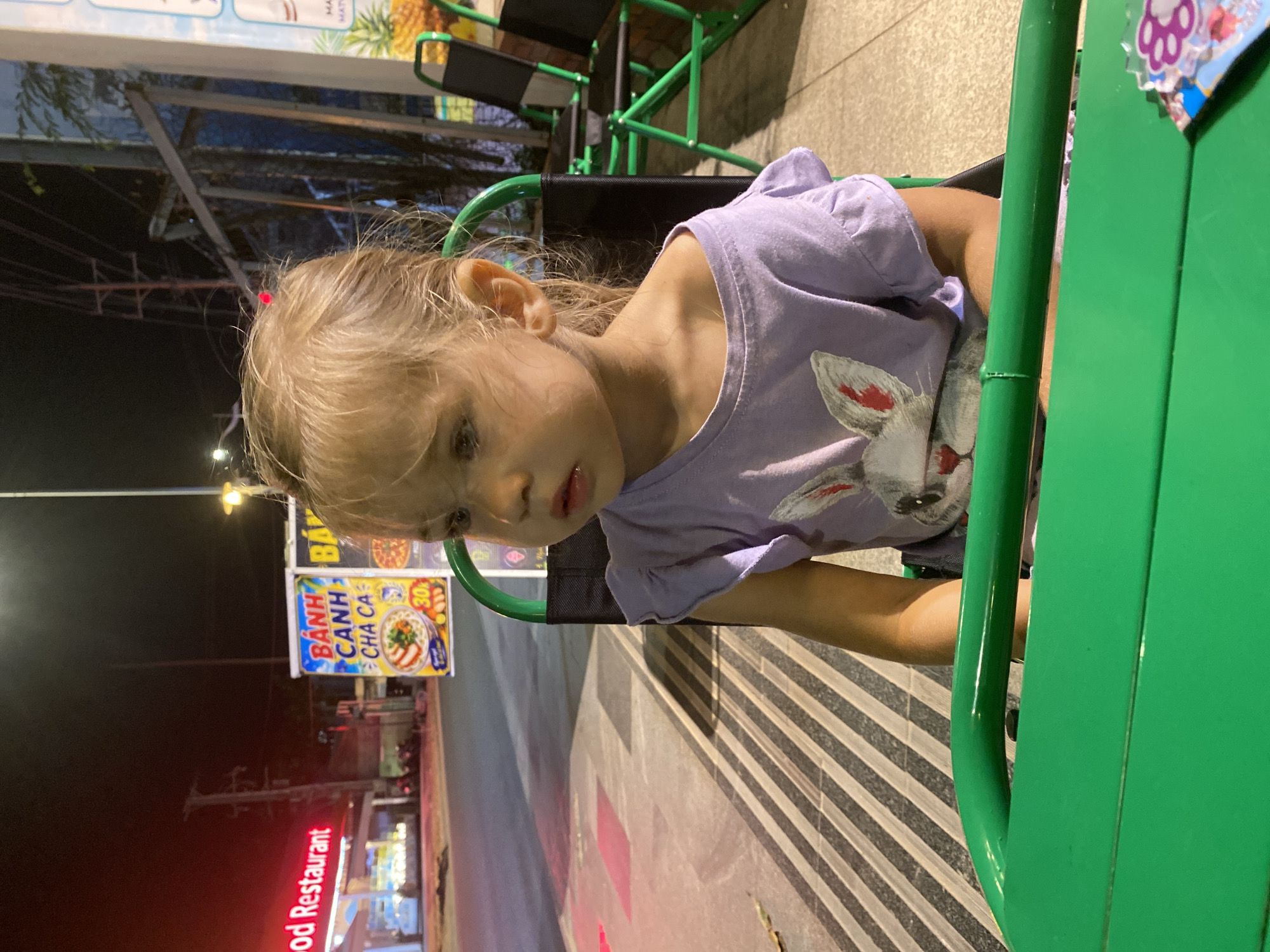 The image shows a young girl with blonde hair, wearing a light purple t-shirt with a bunny graphic, sitting in a green chair. Her head is tilted and she is looking down. The background appears to be outdoors at night, with signs of a street or marketplace visible. One sign, in Vietnamese, displays "Banh Canh Cha Ca" and food imagery. Another sign further in the background reads "Seafood Restaurant" in red neon letters. The overall setting suggests a casual, possibly food-related environment in Phu Thuy Ward, Vietnam, during the evening or night.