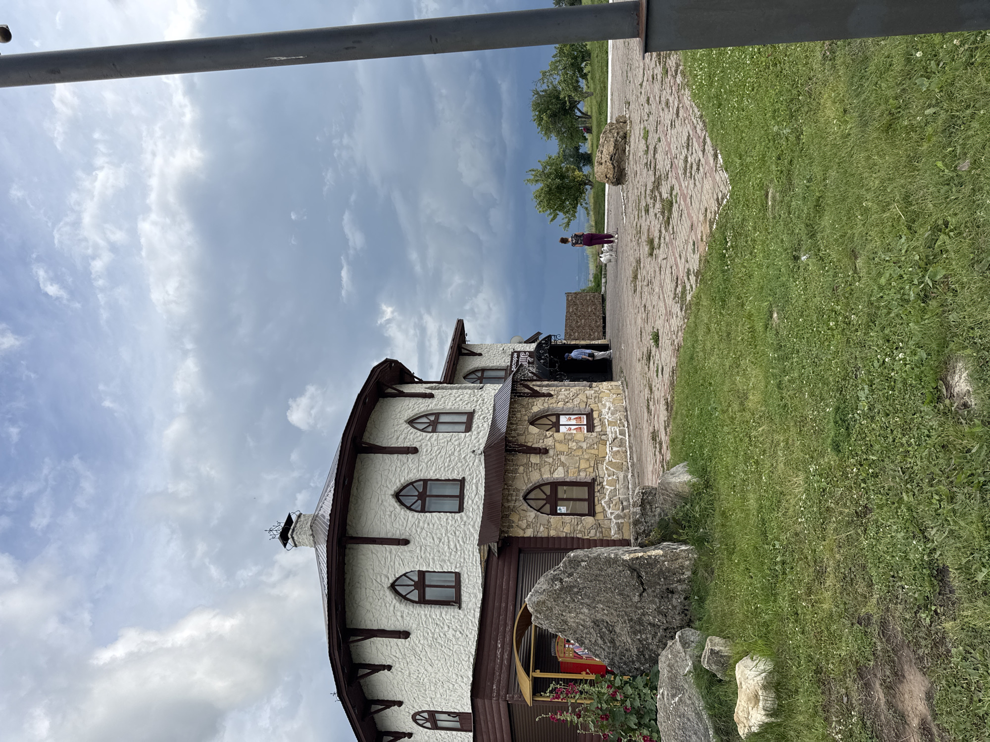 The image shows a circular building with a light-colored, textured exterior and a dark brown, sloped roof. The building has arched windows with dark brown frames. The lower portion of the building is constructed with stone blocks. A large, rough-hewn gray boulder is situated in the foreground, to the left of the building. To the right of the building, a paved area with a reddish-brown brick pattern is visible, with patches of green grass growing through the cracks. A dark gray pole is prominently located on the right side of the frame, partially obscuring the view. In the middle ground, two people are visible. One person, wearing a dark top and purple pants, is standing and appears to be interacting with two small white dogs. Another person, wearing a light-colored shirt and pants, is walking towards the building. The background features a line of green trees and a vast sky filled with dramatic, dark clouds. The scene is located in Yelabuga, Russia, at the I.I. Shishkin Art Gallery and Museum, specifically in the courtyard area.