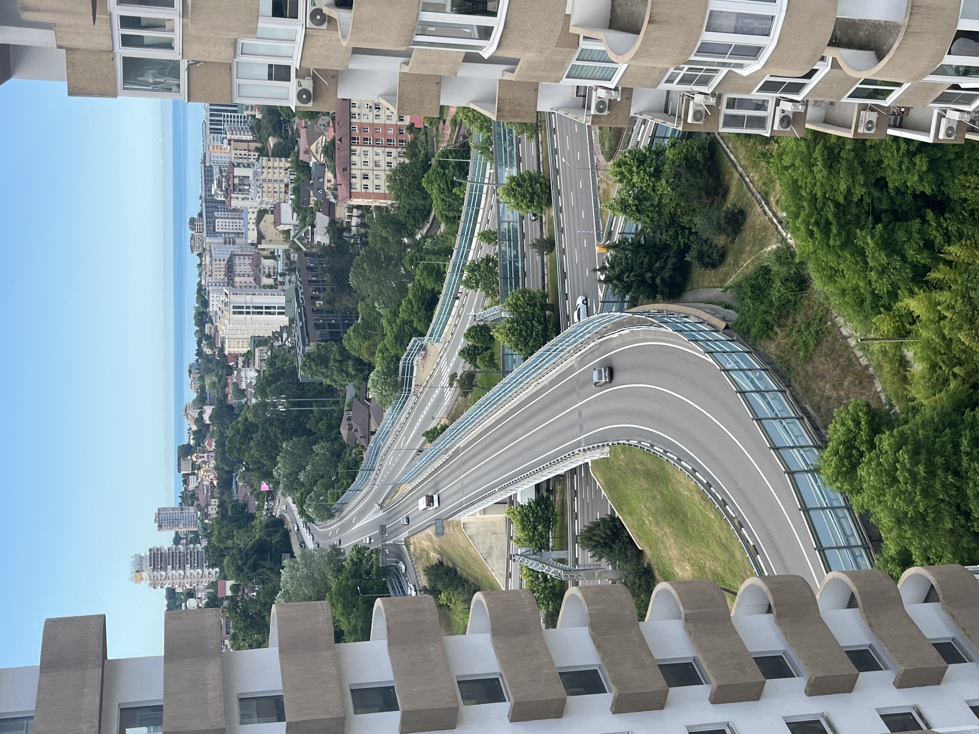 This is an aerial, eye-level, medium shot of the highway system in Sochi, Russia, on a clear day. The highway curves through a lush, green landscape, with trees and vegetation lining the road and surrounding hills. In the distance, the city skyline is visible, with a mix of modern high-rises and older buildings, and the Black Sea stretches to the horizon. Two residential buildings, one on the left and one on the right, frame the scene. The building on the left is a white, multi-story structure with curved balconies. The building on the right is a tan, multi-story structure with curved balconies and numerous windows. Several cars and a bus can be seen driving on the highway. The sky is a clear, bright blue.