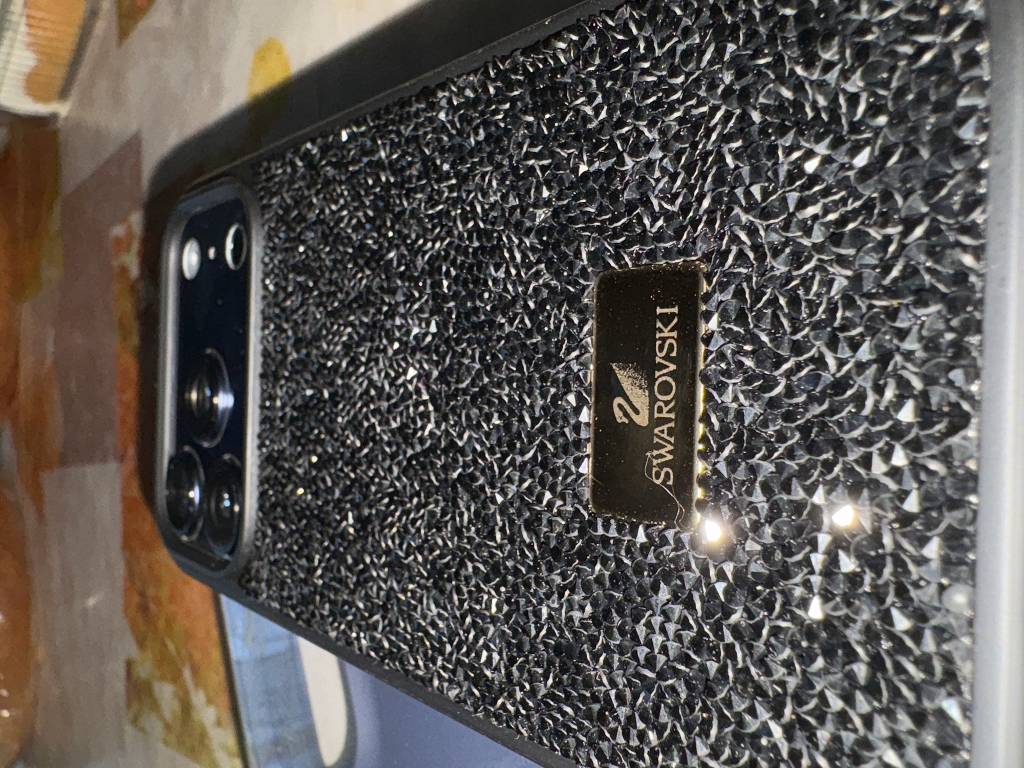 The image shows a smartphone in a black Swarovski crystal-studded case. The case has a rectangular gold plate with the Swarovski logo. The phone's camera array is visible on the left side. The background is blurry, with hints of brown and yellow. The location is not discernible from the image.