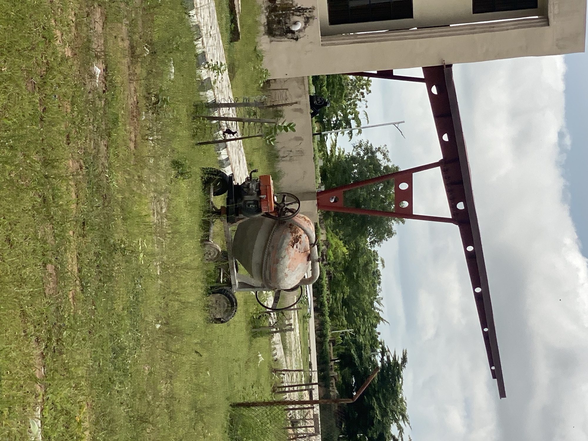 In Jalingo, Nigeria, a cement mixer sits on a grassy patch beside a paved walkway. The mixer, a well-used piece of construction equipment, shows signs of rust and wear. Behind it, a person dressed in black is seated on a low wall, facing away from the camera. In the background, a modern building with large windows stands to the left, and a series of red metal structures with circular cutouts extend outwards overhead. Lush green trees and vegetation are abundant, and a cloudy sky completes the scene.