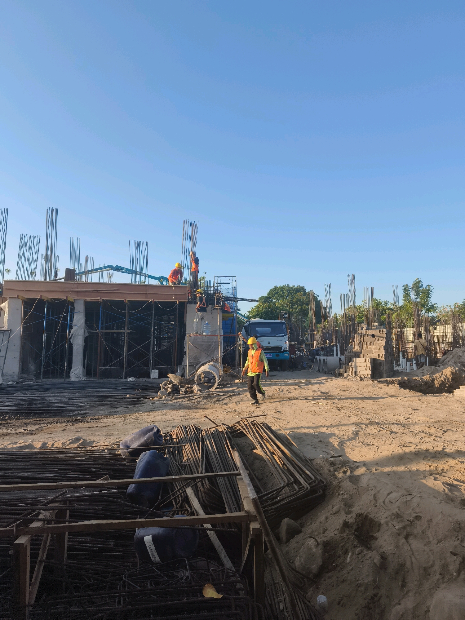 In Denpasar, Indonesia, a construction site is depicted under a clear blue sky. The scene features a multi-story building under construction, characterized by numerous vertical rebar structures protruding from concrete foundations and slabs. Scaffolding and formwork are visible, indicating ongoing construction activities. Several construction workers, distinguished by their yellow hard hats and high-visibility vests (primarily orange), are engaged in various tasks on the elevated levels of the building. One worker, wearing an orange vest and dark pants, is walking across the sandy ground in the foreground, casting a shadow. A concrete mixer truck is parked on the site, positioned near the building. In the immediate foreground, piles of rebar are neatly stacked, alongside some large blue plastic barrels. The environment is a typical construction zone, with exposed earth, sand, and construction materials.