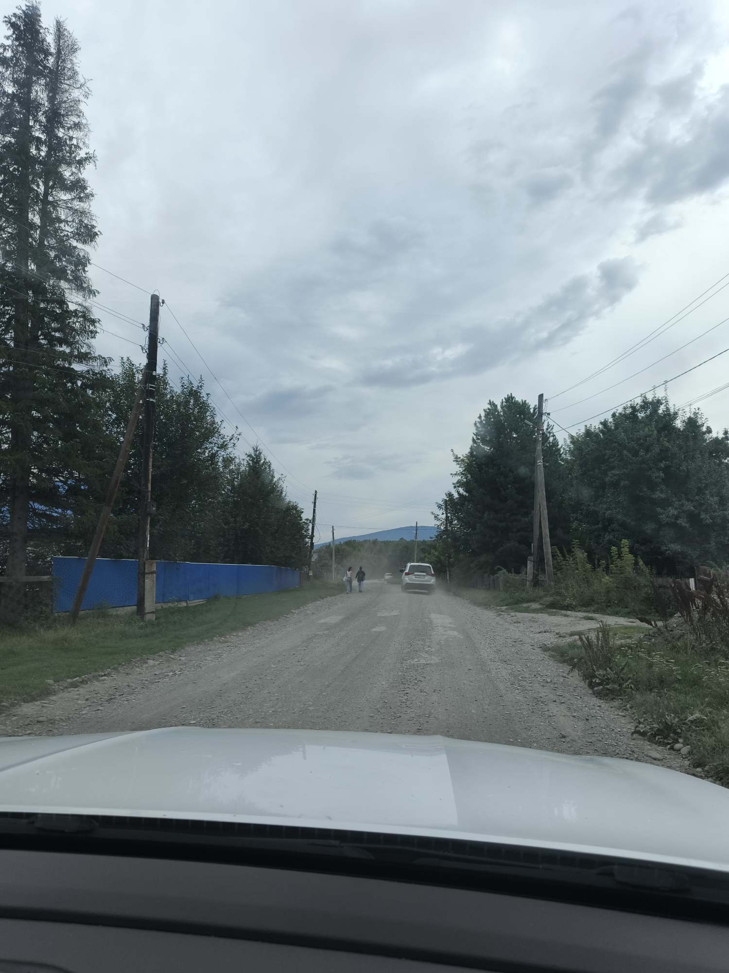 The image captures a view from inside a vehicle looking out at a dusty, unpaved road in a rural setting. On the left side of the road, a long, bright blue fence runs along a grassy area, behind which are dense trees. Several utility poles with wires are visible along the road. Two people are walking away from the camera down the road, and a white SUV is further ahead, also moving away and kicking up dust. The sky is overcast with heavy clouds. In the foreground, the hood of the vehicle the photo is taken from is visible. The overall scene suggests a remote, possibly underdeveloped, location.