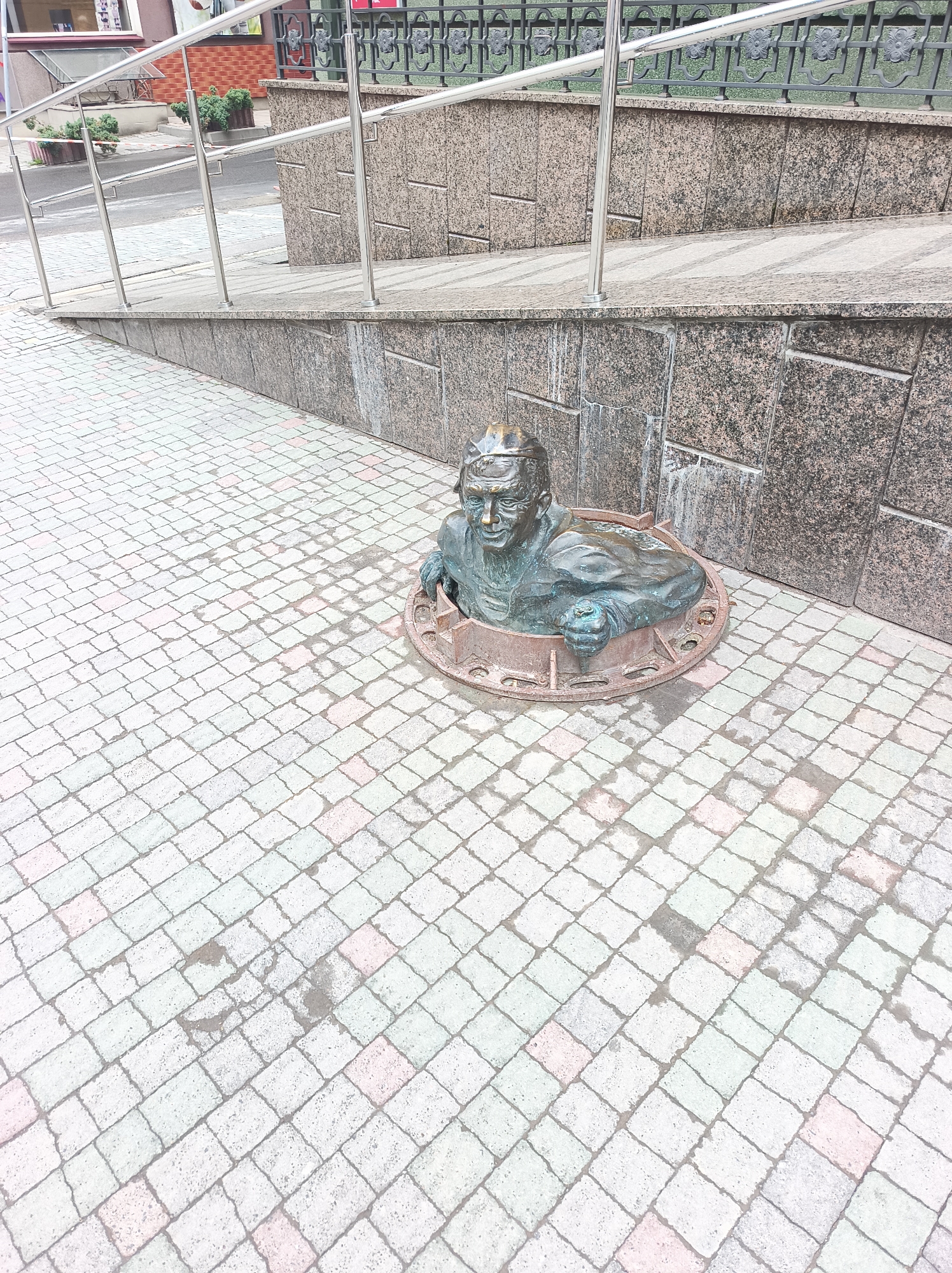 A statue of a man emerging from a manhole cover stands on a tiled sidewalk in Ternopil, Ukraine. The statue depicts a man with glasses wearing a hat or cap, with only his head and shoulders visible above the manhole. The manhole is set into the tiled sidewalk. Behind the statue, there is a granite wall topped with a metal railing. The sidewalk leads to a street and a building in the background.