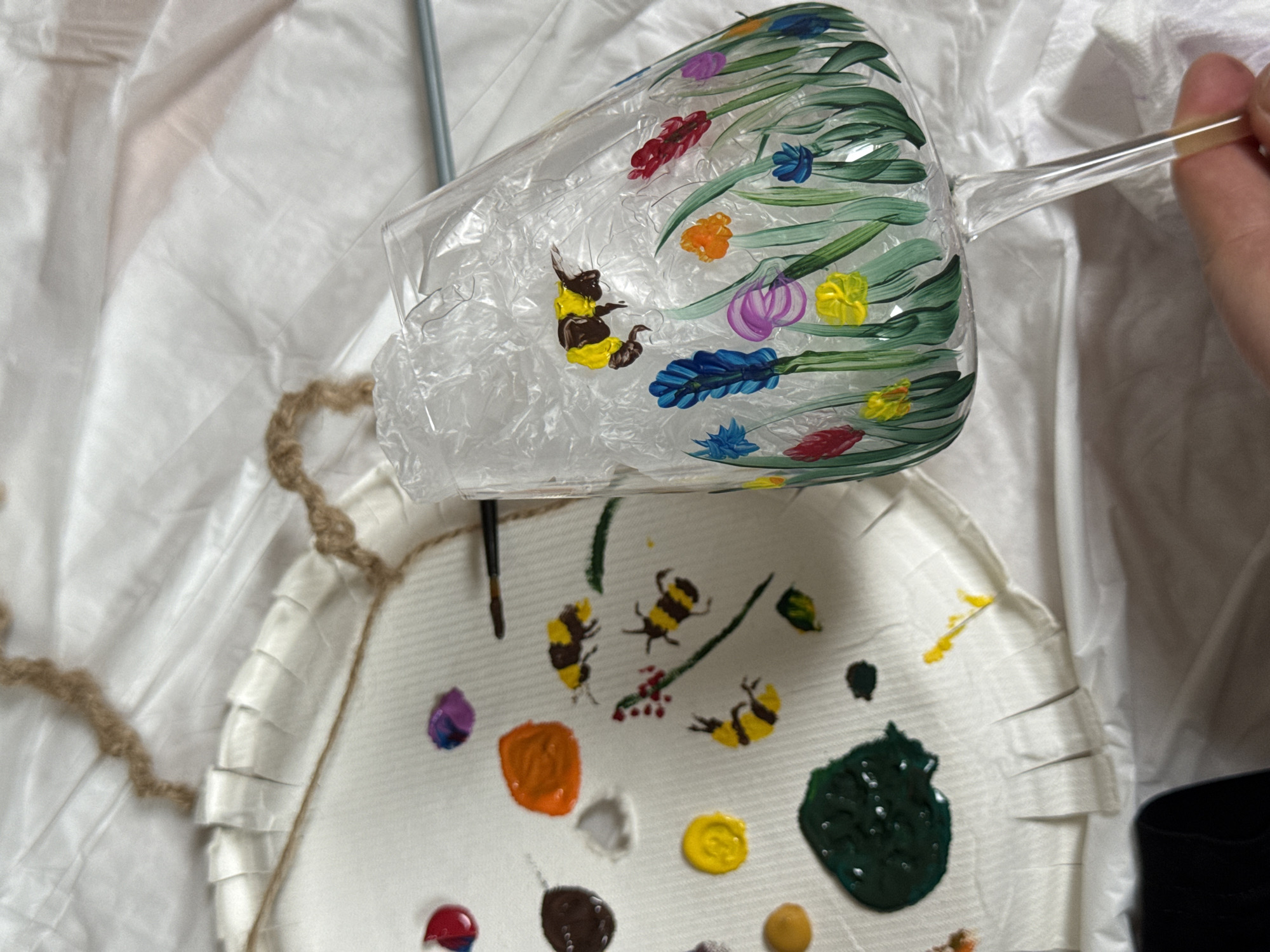 The image shows a clear wine glass being hand-painted with colorful floral and bee designs. The glass is held at an angle over a white paper plate that is being used as a palette. On the palette, there are various small blobs of different colored paints, including red, orange, yellow, brown, green, purple, and white. There are also several bee paintings and streaks of green, likely depicting leaves or stems. The background is a crinkled white plastic sheet, and a thin metal rod is partially visible. Part of a hand holds the wine glass, indicating that someone is in the process of painting it. The image depicts an art project in progress. There is no evidence of the location being Sapronovo, Russia.