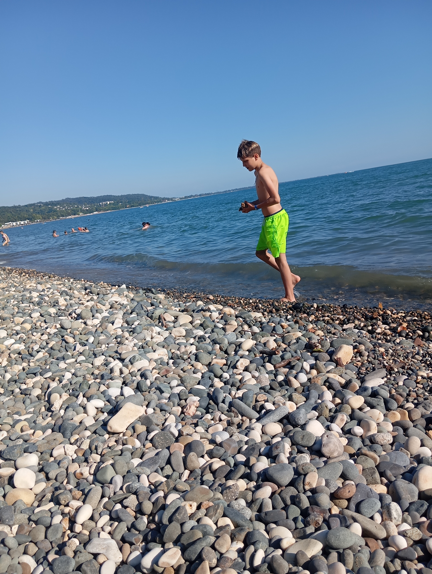 A teenage boy wearing bright green swim trunks stands on a pebble beach near the water's edge, looking down at something in his hands. The sea is calm and blue, with a distant shoreline visible featuring lush green hills and buildings. Several other people are in the water, some swimming and others relaxing. The sky is clear and blue, indicating a sunny day. The beach is covered with a variety of smooth, grey and white pebbles. The overall scene is one of a relaxed summer day at the beach in Sokhumi, Abkhazia.