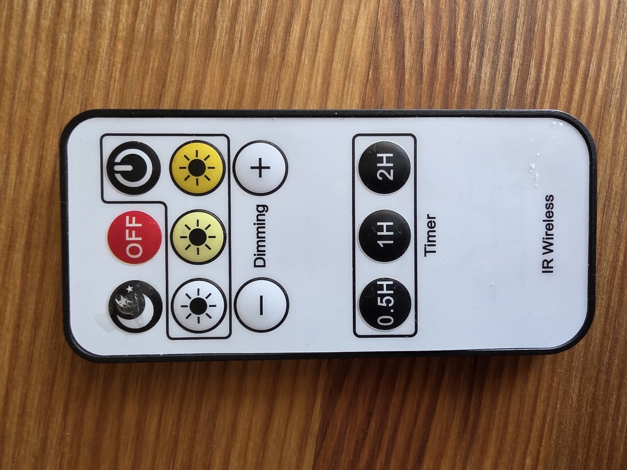 This is a top-down, close-up shot of a white IR wireless remote control lying on a wooden surface. The remote has a black border and a rectangular shape. On the remote, there are several buttons with various symbols and text.
On the left side of the remote, there are four circular buttons arranged vertically. The topmost button features a power symbol (a circle with a line inside). Below it, there's a yellow button with a sun symbol and radiating lines, indicating brightness. The third button is red with the text "OFF" in white. The bottommost button displays a crescent moon with stars and a sun, suggesting a mode or color change.
To the right of these buttons, there are three white circular buttons with symbols. The top one has a plus sign (+), the middle one has a sun symbol, and the bottom one has a minus sign (-). Vertical text labels "Dimming" are situated between these two columns of buttons.
Further to the right, there are three black circular buttons arranged vertically, each containing white text: "2H" at the top, "1H" in the middle, and "0.5H" at the bottom. Vertical text labels "Timer" are next to these buttons.
On the far right of the remote, vertical text reads "IR Wireless". The wooden surface beneath the remote shows a prominent wood grain pattern. No people, specific time of day, weather, or emotional cues are discernible from the image. The context of Omsk, Russia, is provided externally and is not visible within the image itself.