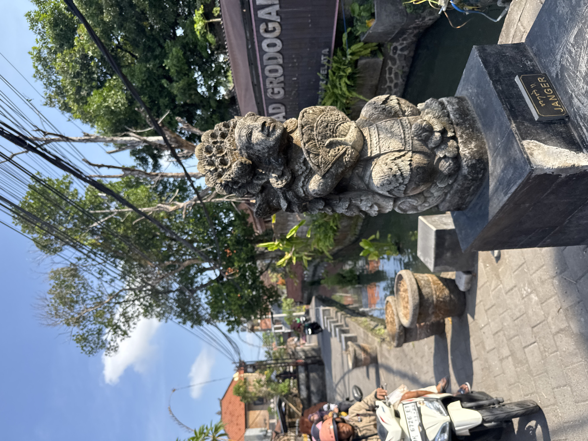 In Denpasar, Indonesia, a stone statue of a woman holding a fan stands on a pedestal in the foreground, inscribed with the word "JANGR". To the left, a person on a white scooter with license plate DK 6726 ET heads away from the camera on a brick pathway. Behind them, a narrow canal with flowing water runs alongside the pathway, bordered by concrete planters. In the background, lush green trees and Balinese-style buildings with orange tiled roofs line the street, which has utility poles and wires overhead. A sign that reads "AD GRODOGAN" is visible on a building in the distance. The sky is blue with scattered white clouds, and the sun casts shadows on the scene.