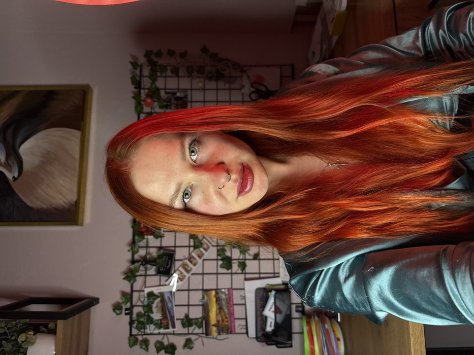 A fair-skinned woman with vivid red-orange hair and green eyes poses at a slightly tilted angle. She has a nose ring and wears a teal silky shirt. Behind her is a black wire grid with ivy and pictures attached. To her left is a painting of an animal, possibly a bird, in a gold frame. Underneath the painting is a small shelf with a plant. The lighting gives a red tint to her face, and there is a red light fixture at the top of the frame.