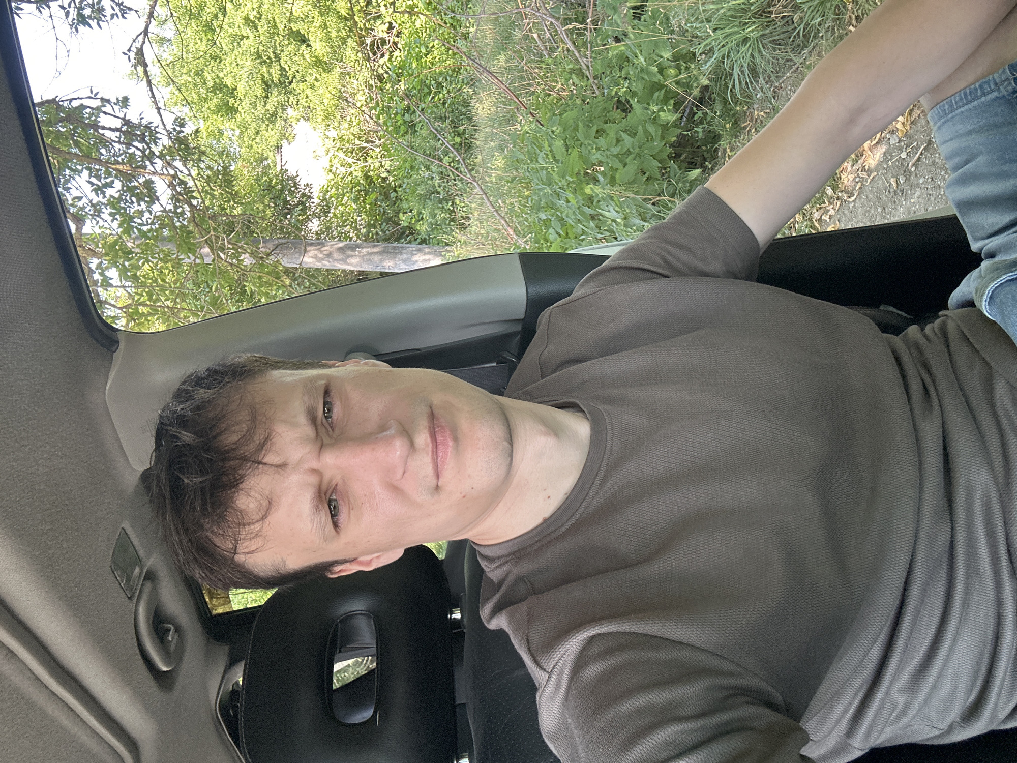 A man with short brown hair and fair skin is seated in the driver's seat of a car, taking a selfie. He is wearing a brown t-shirt and denim shorts. The car's interior is visible, including the headrest of the passenger seat and the car's ceiling. Outside the car window, there is lush green foliage, trees, and a dirt path, suggesting a rural or natural setting. The lighting indicates it is daytime, likely sunny. The man's facial expression is neutral, with a slight frown. The specific location in Makiv, Ukraine is not discernible from the visual information in the image.