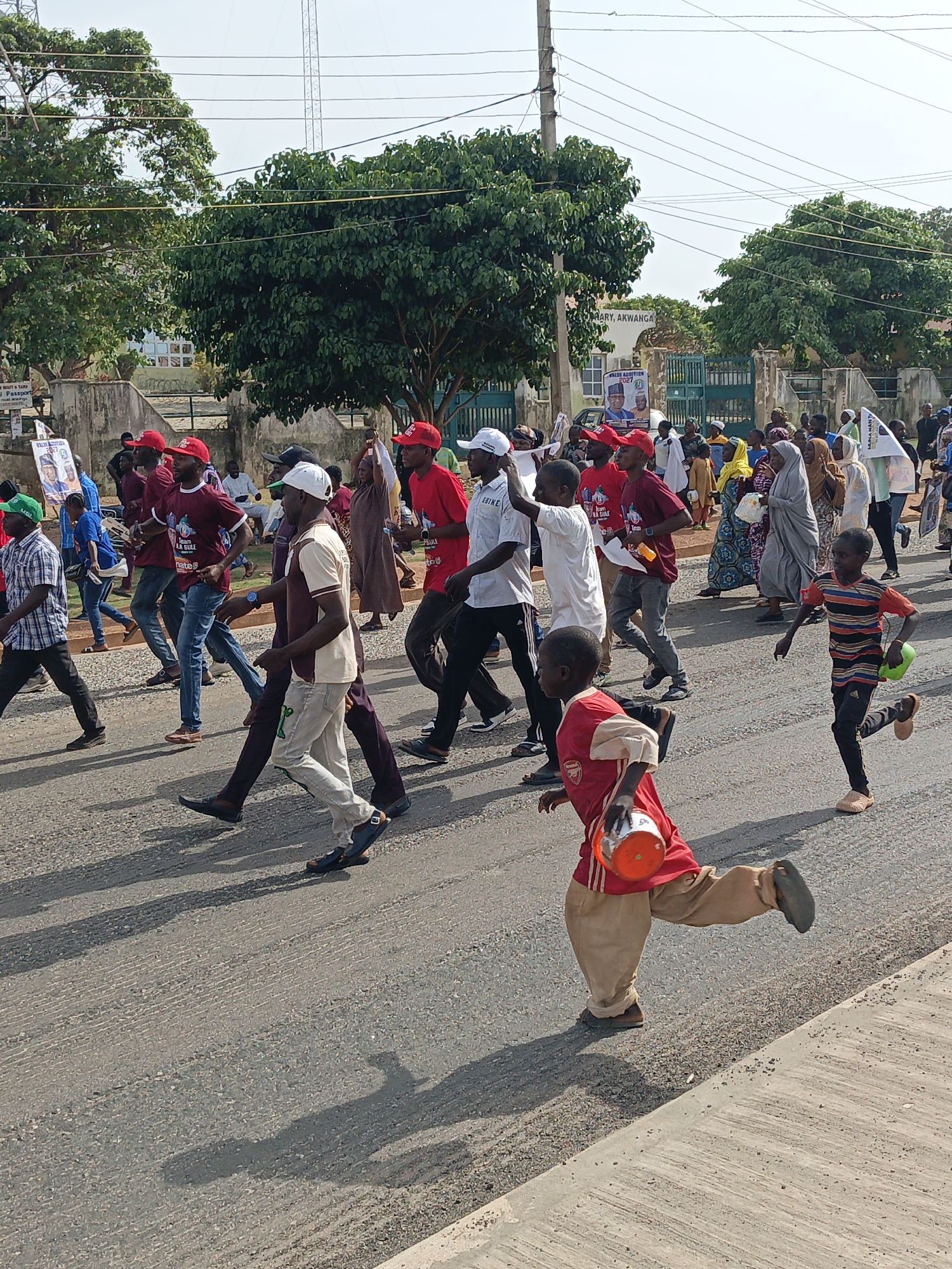 A crowd of people is walking along a street in Akwanga, Nigeria. Most appear to be marching, and some wear red shirts and caps. Two children are running along the road in the foreground. One child is wearing a red Arsenal t-shirt and beige pants, carrying an orange pot. The other child wears a striped shirt and black pants, carrying a small green ball. A building in the background has signage: "PRIMARY, AKWANGA" and "VALUE ADDITION 2027". A political poster featuring two men, with text that reads "Passport", "Team LA SULE", and "SULE". Overhead power lines are visible.