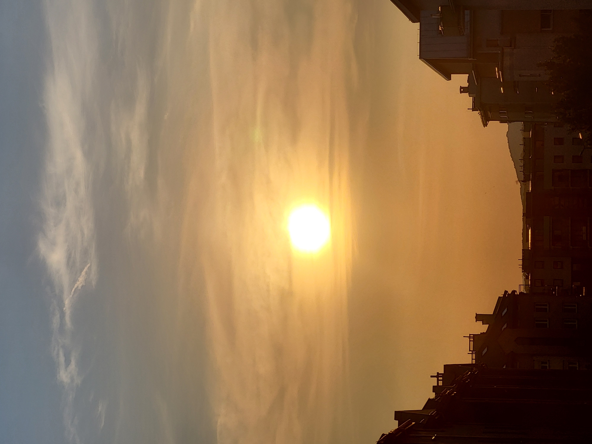 This is a vertical, eye-level, outdoor shot of a sunset over a city. The sun is a bright, white-yellow orb, situated in the center of the frame, surrounded by a hazy orange sky. The sky transitions to a pale blue at the very top of the image. Several wispy clouds are visible, some catching the sunlight and appearing tinged with yellow and pink. The foreground is dominated by the silhouettes of apartment buildings on either side of the frame. The buildings on the left are darker and appear to be older, with visible rooftops and windows. The buildings on the right are lighter in color and appear more modern, with angled roofs and balconies. In the far distance, a hazy, dark silhouette of a hill or mountain range can be seen against the sunset. There are no people visible in the image. The overall impression is one of a calm, urban evening.