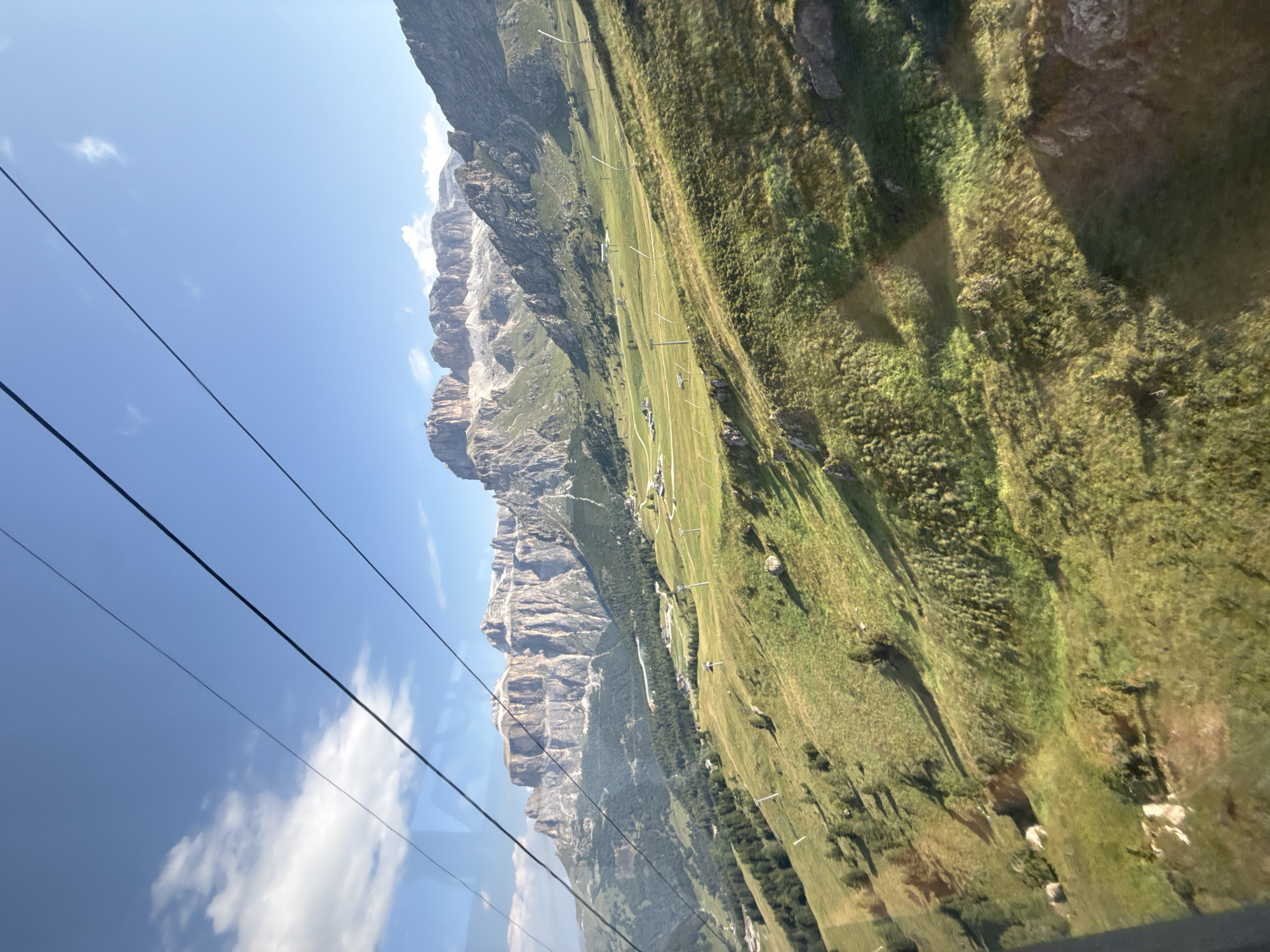 The image shows a scenic mountain landscape, likely taken from a cable car, overlooking the Sella group in the Dolomites near Canazei, Italy. The foreground is dominated by a lush, green, sloping meadow dotted with scattered rocks and sparse trees. The meadow is crisscrossed with subtle paths or ski tracks, and several ski lift pylons are visible, indicating a mountain resort area. In the midground, more trees form a denser forest, and small clusters of buildings can be seen nestled within the valley. The background features the imposing, jagged peaks of the Sella group, bathed in sunlight under a clear blue sky with scattered white clouds. Three thick black cables, presumably part of the cable car system, are prominently stretched across the upper portion of the frame, suggesting the viewer's vantage point. The lighting suggests it is daytime, likely late morning or early afternoon, with strong shadows cast across the landscape. There are no people or animals visible in the image.