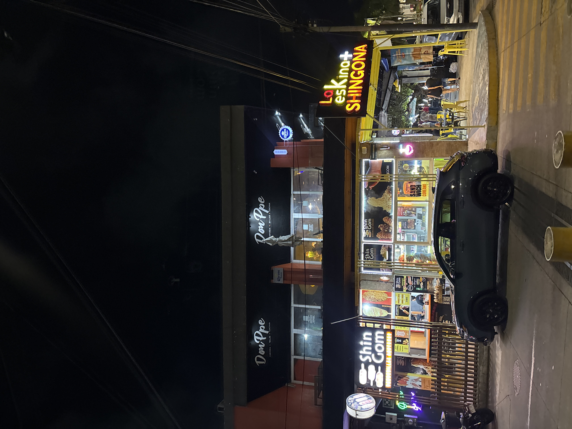 Here's a detailed description of the image:
**Overall Scene:** The image shows a nighttime street scene with multiple businesses in Veracruz City, Mexico. The perspective appears to be from street level, looking toward the establishments.
**Buildings and Businesses:**
* **Don Ppe:** A building with the name "Don Ppe" prominently displayed is visible. The text indicates it's a bar and restaurant. There is a Corona beer sign attached to the wall.
* **La Eskina+ SHINGONA:** A sign reads "La Eskina+ SHINGONA" on top of the building that seems to house more businesses. This appears to be a restaurant or food vendor.
* **Shin Gom:** A business with a lot of signage, appearing to specialize in Korean food, specifically corndogs and sandwiches.
**Vehicles and People:**
* A dark-colored car is parked alongside the building and partially on the street.
* People are visible within the "La Eskina+ SHINGONA" establishment, seated at tables, suggesting the business is open and serving customers.
**Street and Environment:**
* The street is paved and marked with yellow stripes. There are sidewalk barriers.
* Power lines are strung overhead, typical of an urban environment.
* The lighting is artificial, from streetlights and business signs, contributing to the nighttime atmosphere.
**Other Details:**
* The image has been rotated or was taken in a vertical orientation.
* The perspective is slightly tilted.
**Location:** Veracruz City, Mexico. This is inferred from the text on the businesses and the general urban environment depicted.