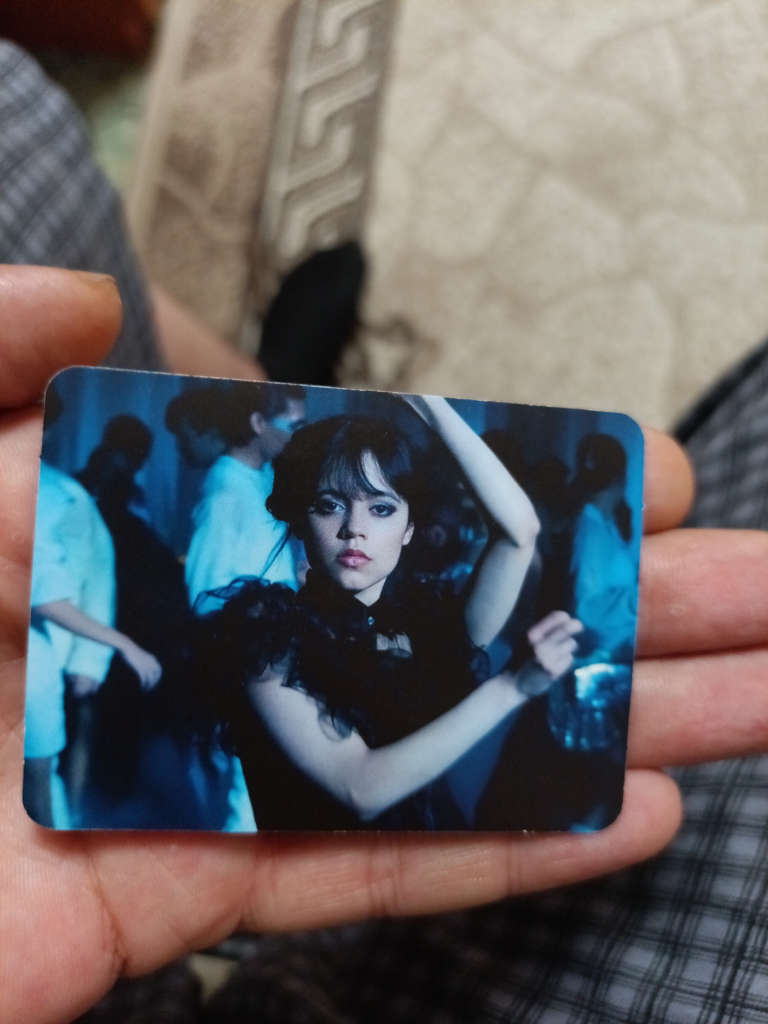 An image card, held by a person, features a still from the TV show "Wednesday". The card depicts the character Wednesday Addams, portrayed by Jenna Ortega, in a black dress with ruffled sleeves and collar, her hair styled with bangs. She is positioned in the foreground with her head slightly tilted and a subtle expression. Behind her, blurred figures in a dimly lit setting suggest a party or social gathering, with blue lighting enhancing the atmosphere. The background is dark and indistinct, with some individuals visible in lighter clothing. The card has rounded corners. The image is not located in the city of Nyagan, Russia, as there are no discernible landmarks or contextual clues to suggest this location.