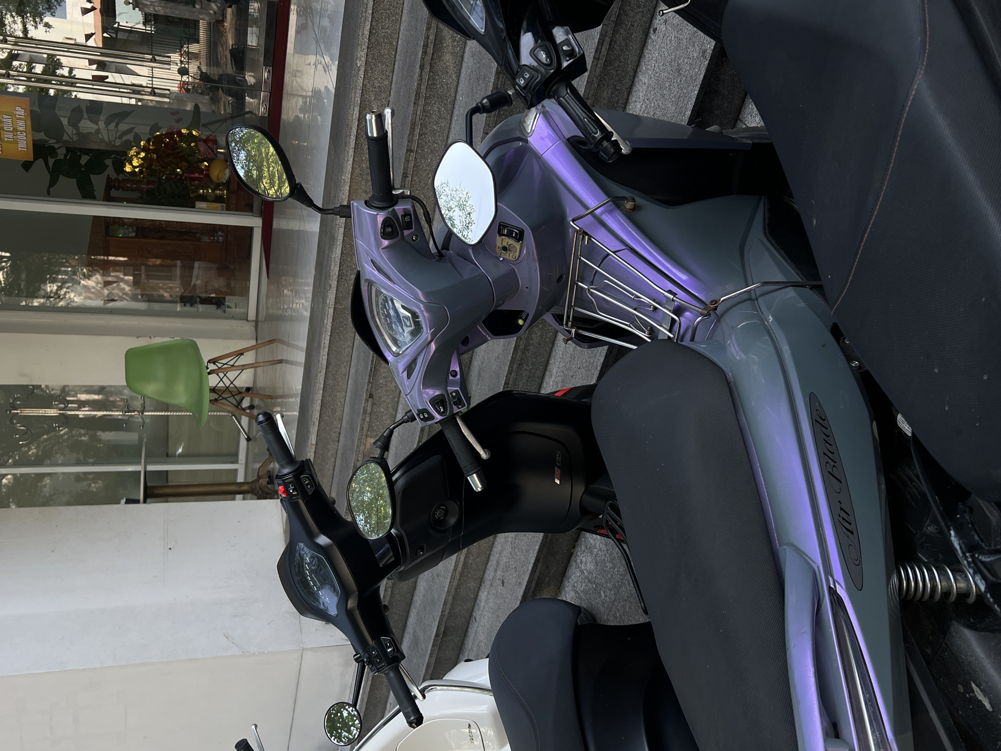 The image shows a lineup of parked motorbikes on a set of concrete stairs. The backdrop includes a building with a glass storefront reflecting foliage and interior elements like a green chair and a wooden table. The location is suggested to be Nam Nha Trang, Vietnam, based on the prompt. A prominent "Air Blade" logo is visible on one of the motorbikes.
