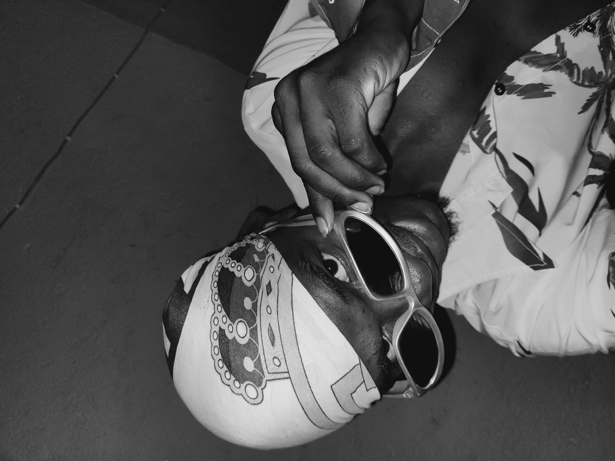 A black and white, close-up, medium shot shows a person wearing a patterned headscarf and large sunglasses. The person is looking down and to the right, with their right hand touching the sunglasses. The person is wearing a short-sleeved collared shirt with a tropical print of leaves and palm trees. The background is dark and appears to be a textured wall. The scene is indoors, possibly in a residence in Bukuru, Nigeria, given the provided information. The lighting is dim, creating strong shadows.