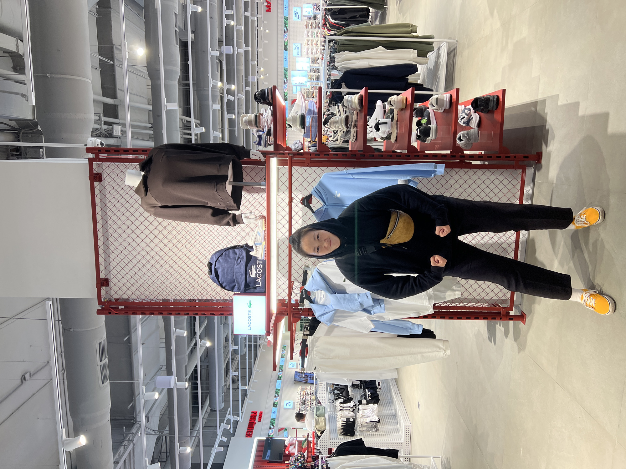 A young person is captured in a bright, modern retail store, posing for the camera with a faint smile. She is wearing a black hooded sweatshirt, black trousers, and distinctive yellow sneakers, complemented by a tan fanny pack worn across her chest. Her stance is casual, with arms bent and fists loosely clenched.
The store features an industrial design with high, exposed ceilings displaying large gray ducts, pipes, and numerous white track lights. The flooring is a light, uniform color. Merchandise is neatly arranged on various display units. Behind the person is a prominent red metal grid structure, from which blue and white apparel hangs. To the left, a mannequin sports a brown jacket. A navy blue Lacoste backpack is displayed on a shelf, next to a clear "LACOSTE" sign. Further in the background, rows of shoes are visible on red display shelves, alongside racks of folded clothing. Visible brand logos in the distance include "Nike" and "Puma," indicating a multi-brand sports or casual wear store. A sign "KEDOBЫШ WOMEN" is also visible. The setting suggests an active shopping environment during the daytime or evening, with ample artificial lighting.