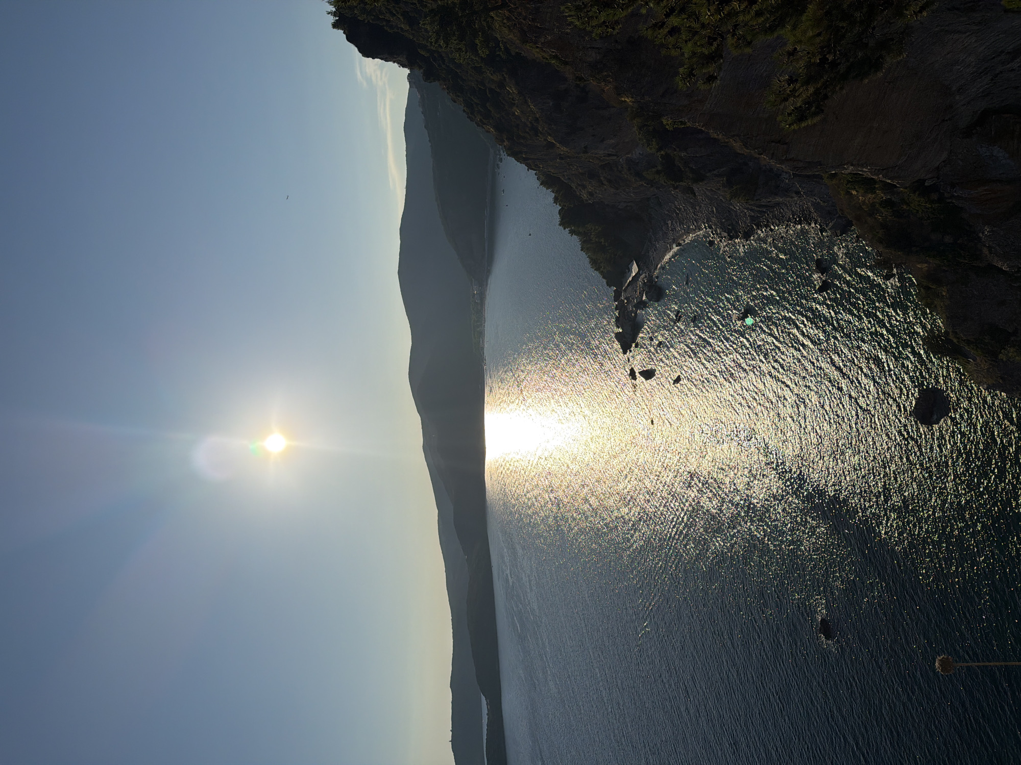 The image is a high-angle shot of a coastal landscape at sunset, likely taken from a vantage point overlooking the Adriatic Sea. The sun, positioned in the upper center of the frame, casts a brilliant, shimmering reflection across the water. Jagged cliffs covered in dark green vegetation dominate the right side of the image, descending towards a rocky shoreline. In the distance, rolling hills form a dark silhouette against the pale blue sky. The water appears deep blue, with visible ripples and a few scattered rocks or small islands near the shore. There are no people or visible structures in the immediate foreground or on the water, apart from what appear to be a few small boats docked or anchored near the coastline in the distance. The overall impression is one of natural beauty and tranquility. Given the description and visual cues, this scene strongly resembles the coastline near Budva, Montenegro, possibly from a viewpoint offering a panoramic vista of the bay and surrounding hills.
