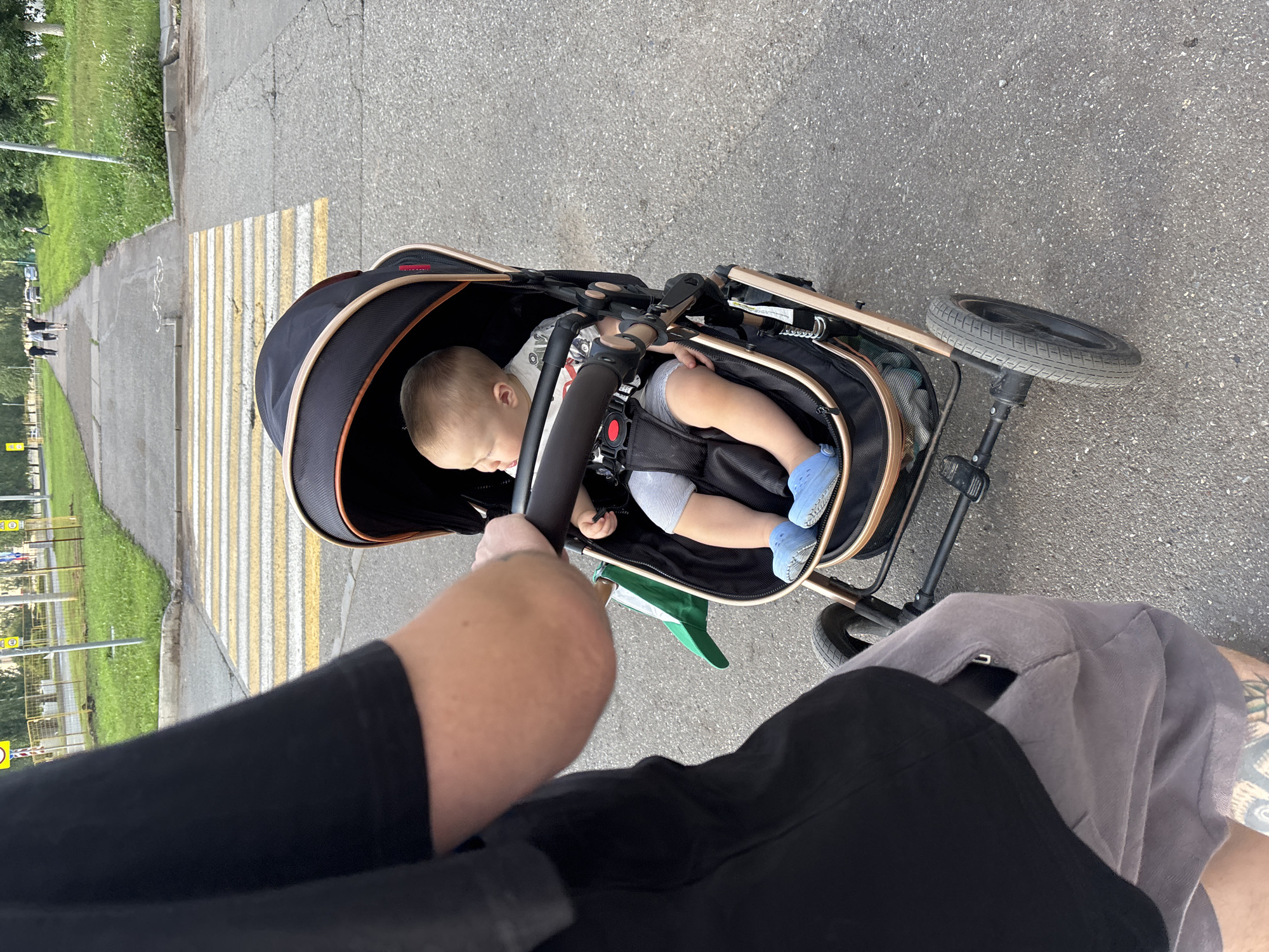 A person wearing a black t-shirt and grey shorts is pushing a baby in a stroller on a paved sidewalk in Naberezhnye Chelny, Russia. The baby, wearing blue shoes and grey shorts, is sitting in the stroller. A pedestrian crossing with yellow and white stripes is visible in the background. The area is lined with grass and trees, suggesting a park or residential area within the city.