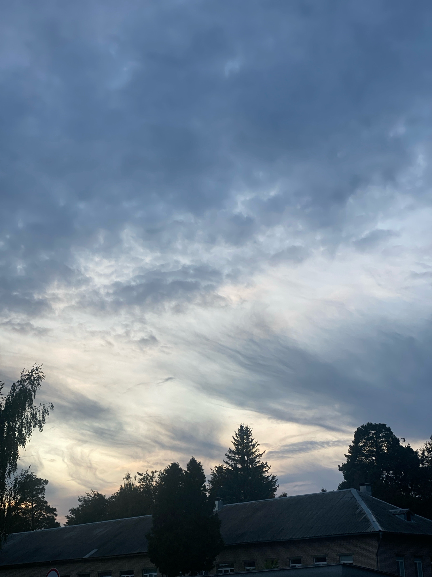 The image depicts a twilight sky over a low-rise brick building with a gray, corrugated roof. Silhouetted trees, including a prominent birch on the left and evergreens on the right and center, are visible against the sky. The sky is filled with textured, wispy clouds, with a lighter area near the horizon suggesting the last vestiges of daylight. A circular red and white sign is partially visible on the left side of the building. The scene appears to be in a semi-urban or suburban area of Machulishchy, Belarus. There are no people or overt actions visible in the image.